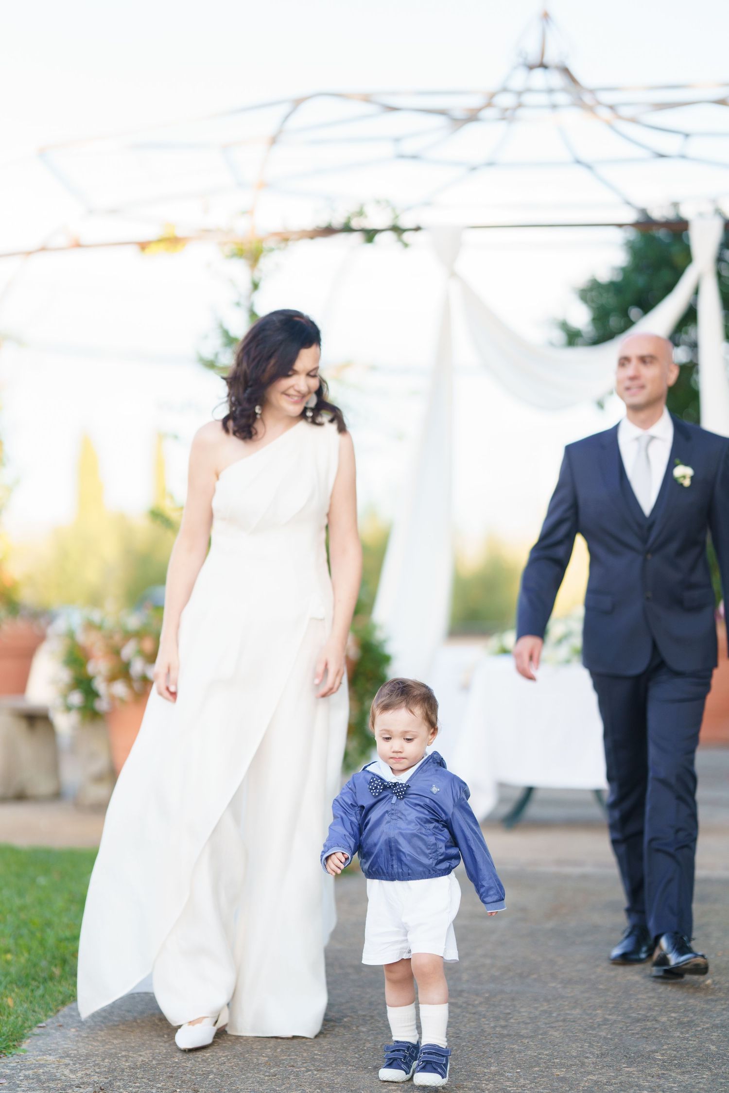 Wedding party strolling on garden pathway with decorative white tent overhead.