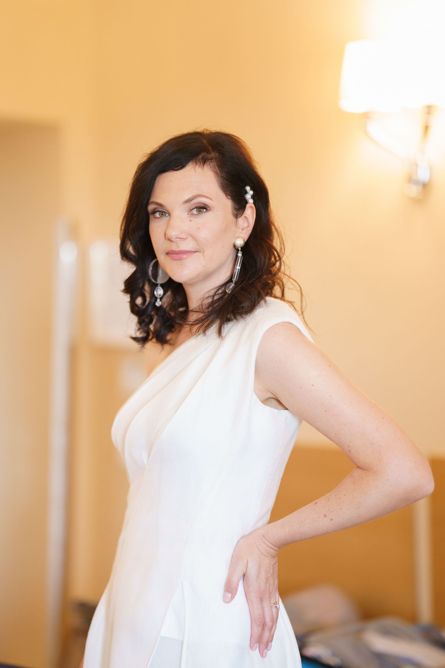 Woman in elegant white dress poses gracefully against cream colored wall with soft lighting.