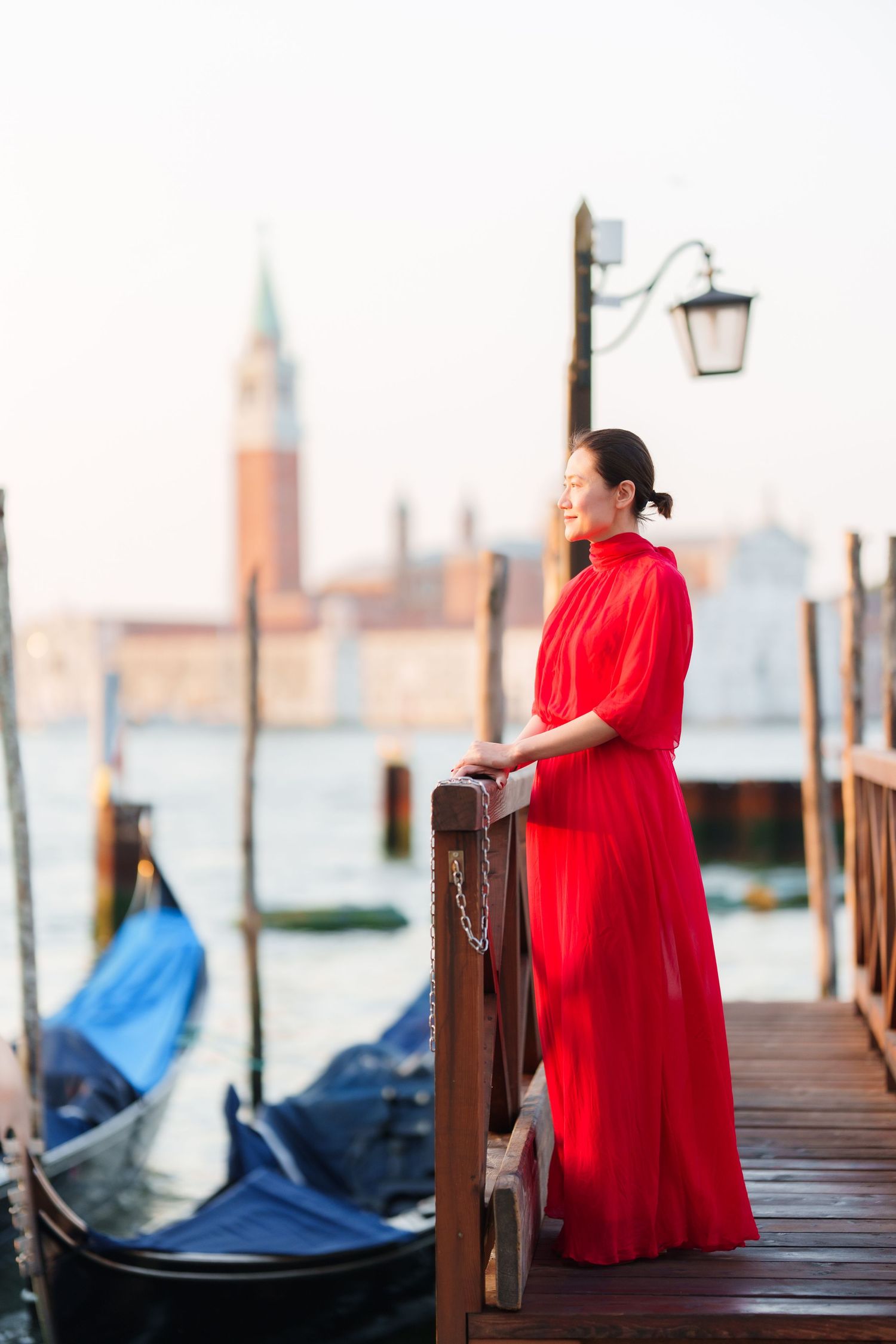 Woman in red dress standing on Venice dock with gondolas and San Giorgio Maggiore church in background.