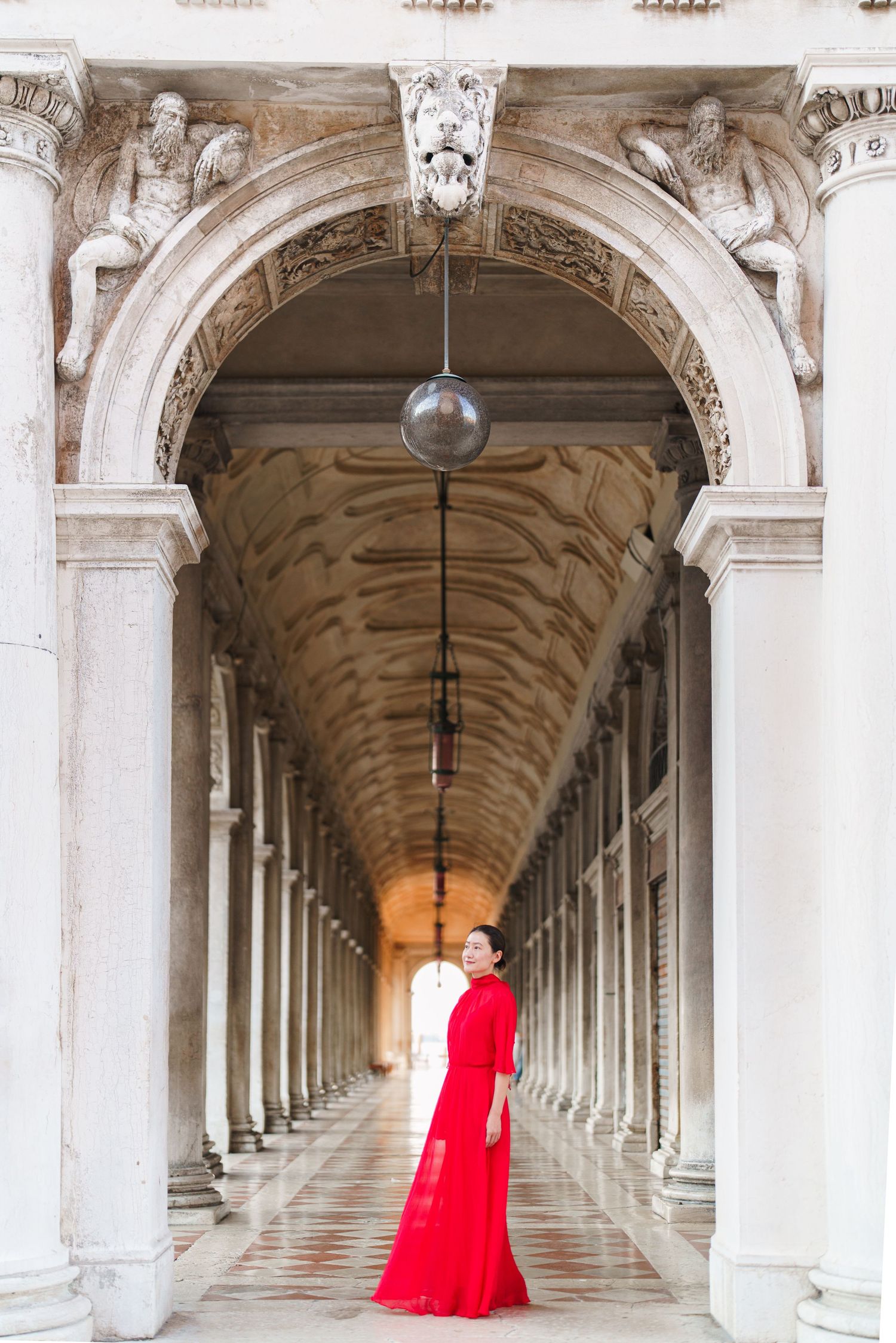 Woman in red gown standing in ornate Venetian arcade colonnade with arched ceiling perspective.