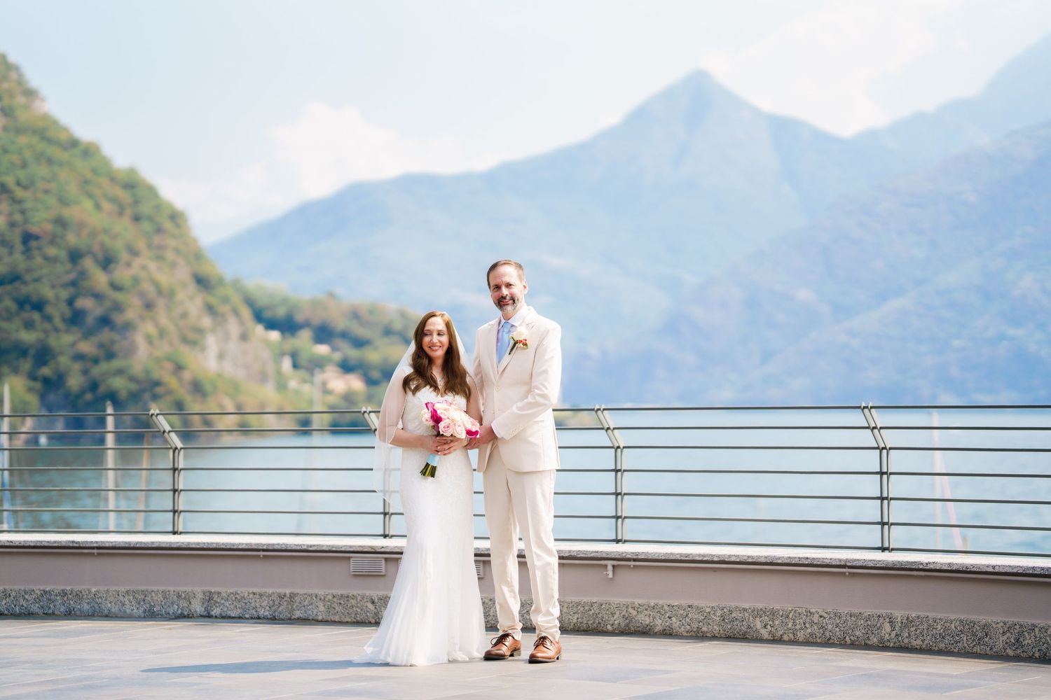 Bride and groom posing on lakeside terrace with stunning mountain and lake views on their wedding day.