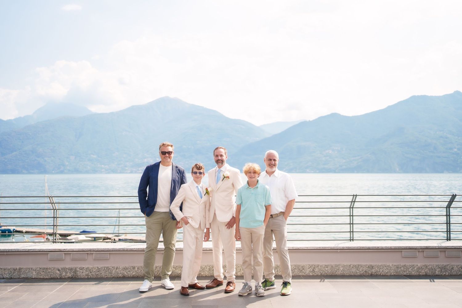 Five men posing together on a lakeside promenade with mountain views and clear skies in the background.