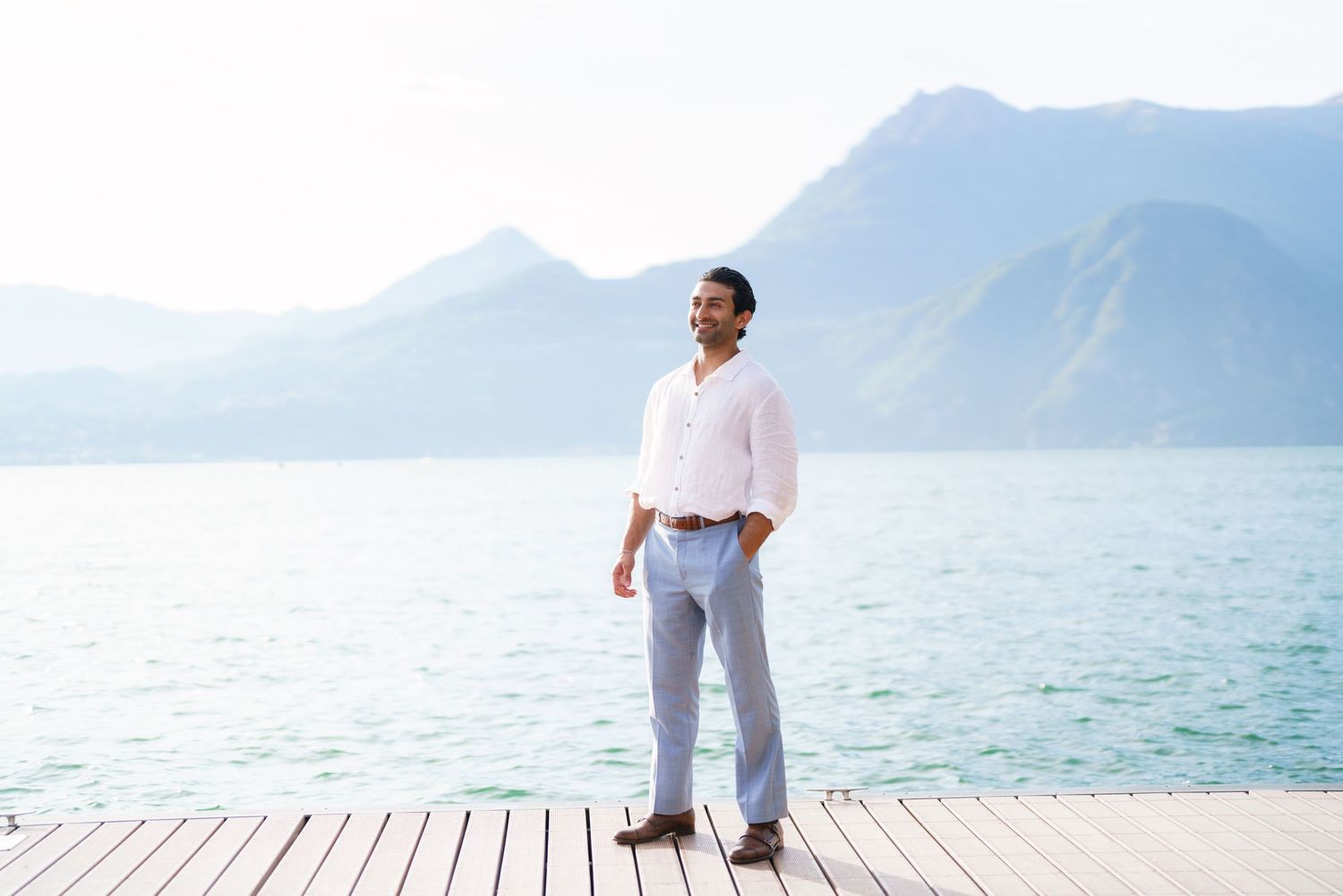 Man in white shirt and light blue pants standing on wooden dock with turquoise lake and mountains behind him.
