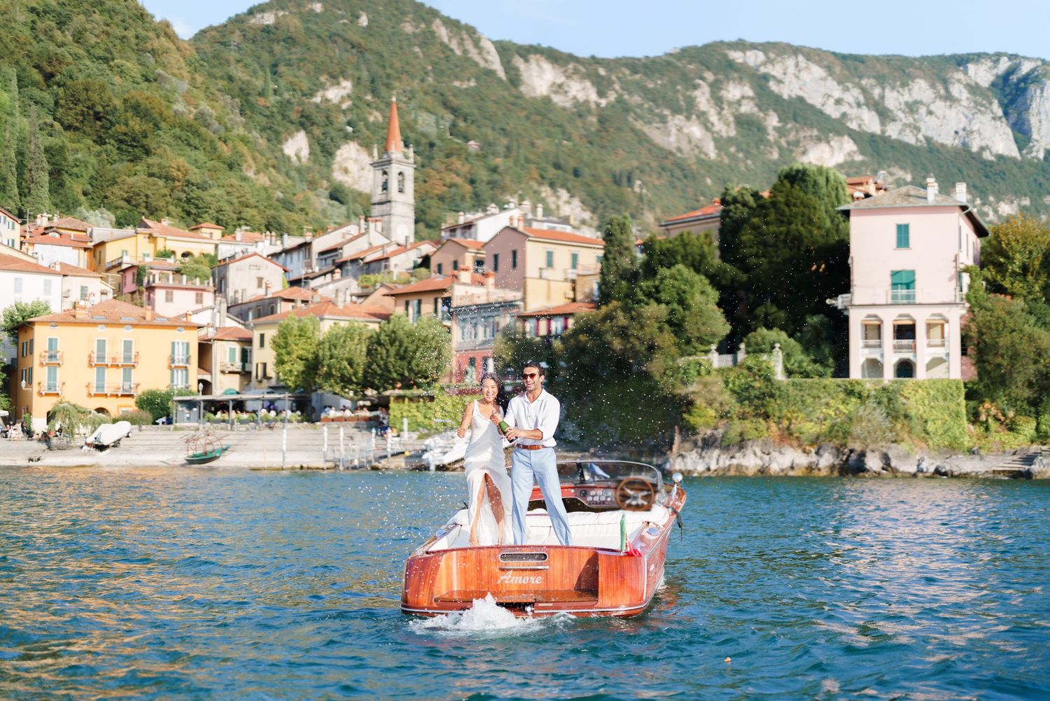 Couple standing on a wooden boat on Lake Como, Italy, with colorful lakeside village and mountains in background.