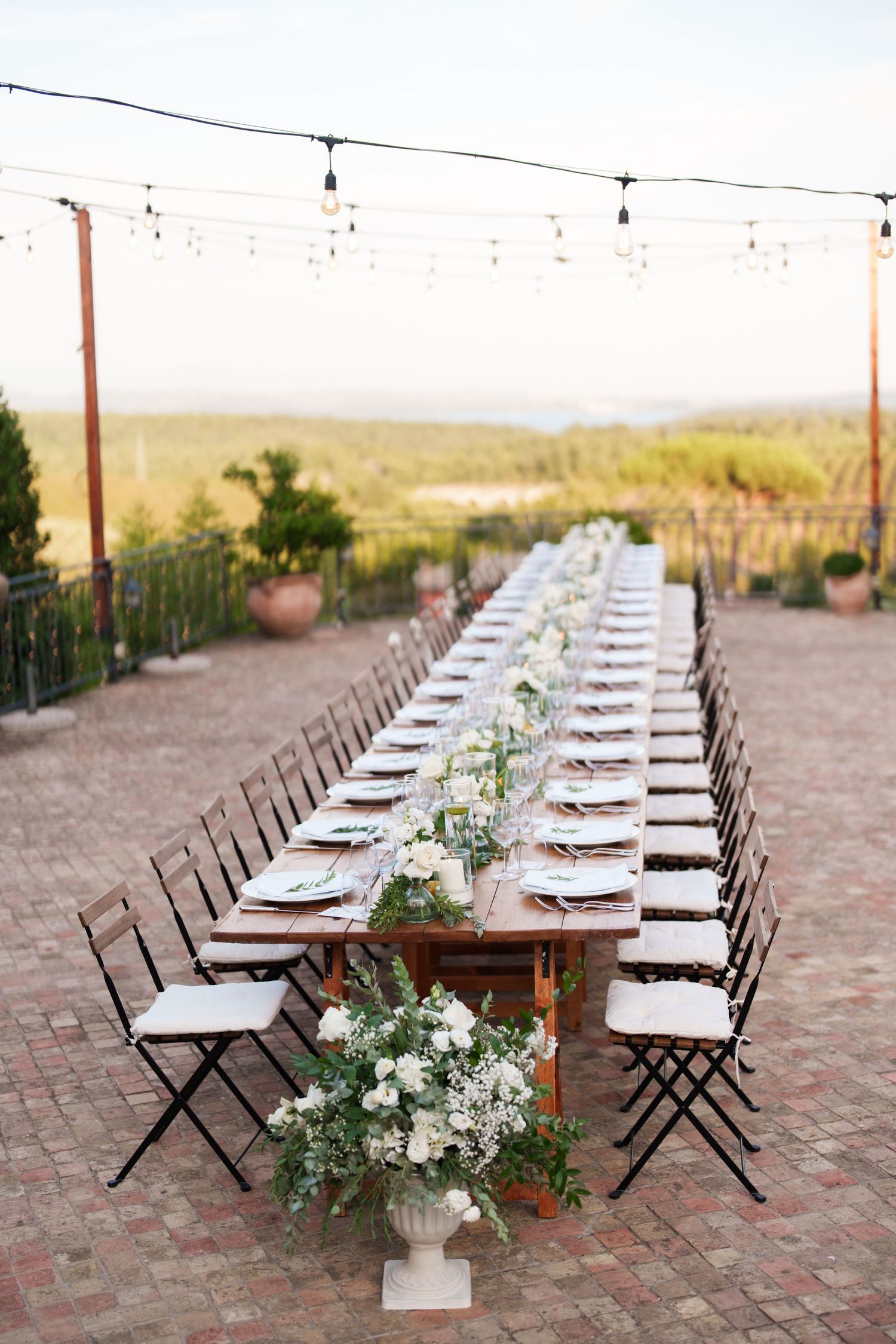 Elegant outdoor wedding reception with long wooden farm table, white floral centerpieces, string lights, and scenic countryside views.