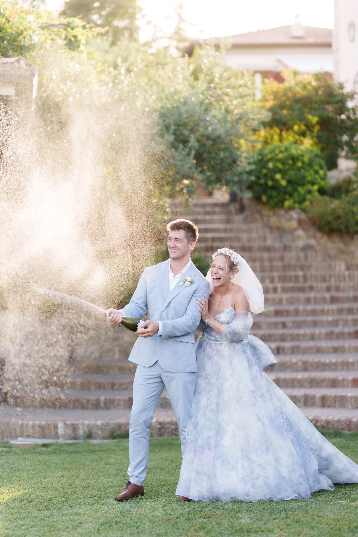 Joyful bride and groom in blue attire popping champagne bottle outdoors on green lawn at wedding celebration.