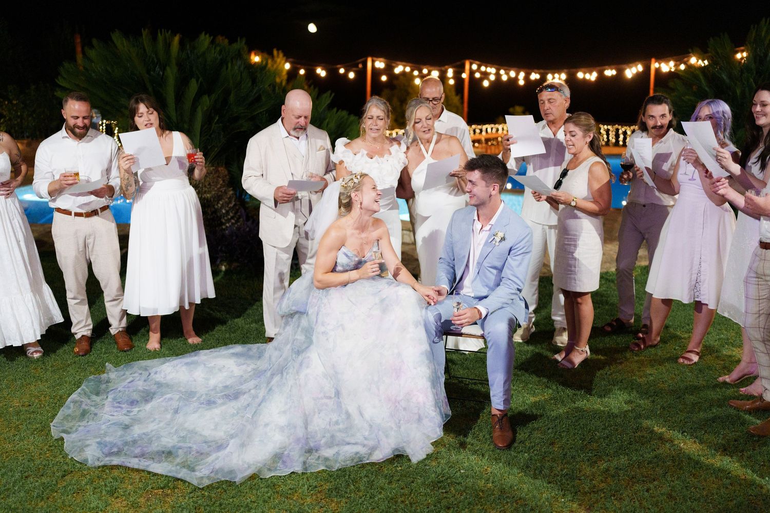 Bride and groom seated outdoors at wedding reception surrounded by guests under string lights at elegant garden ceremony.
