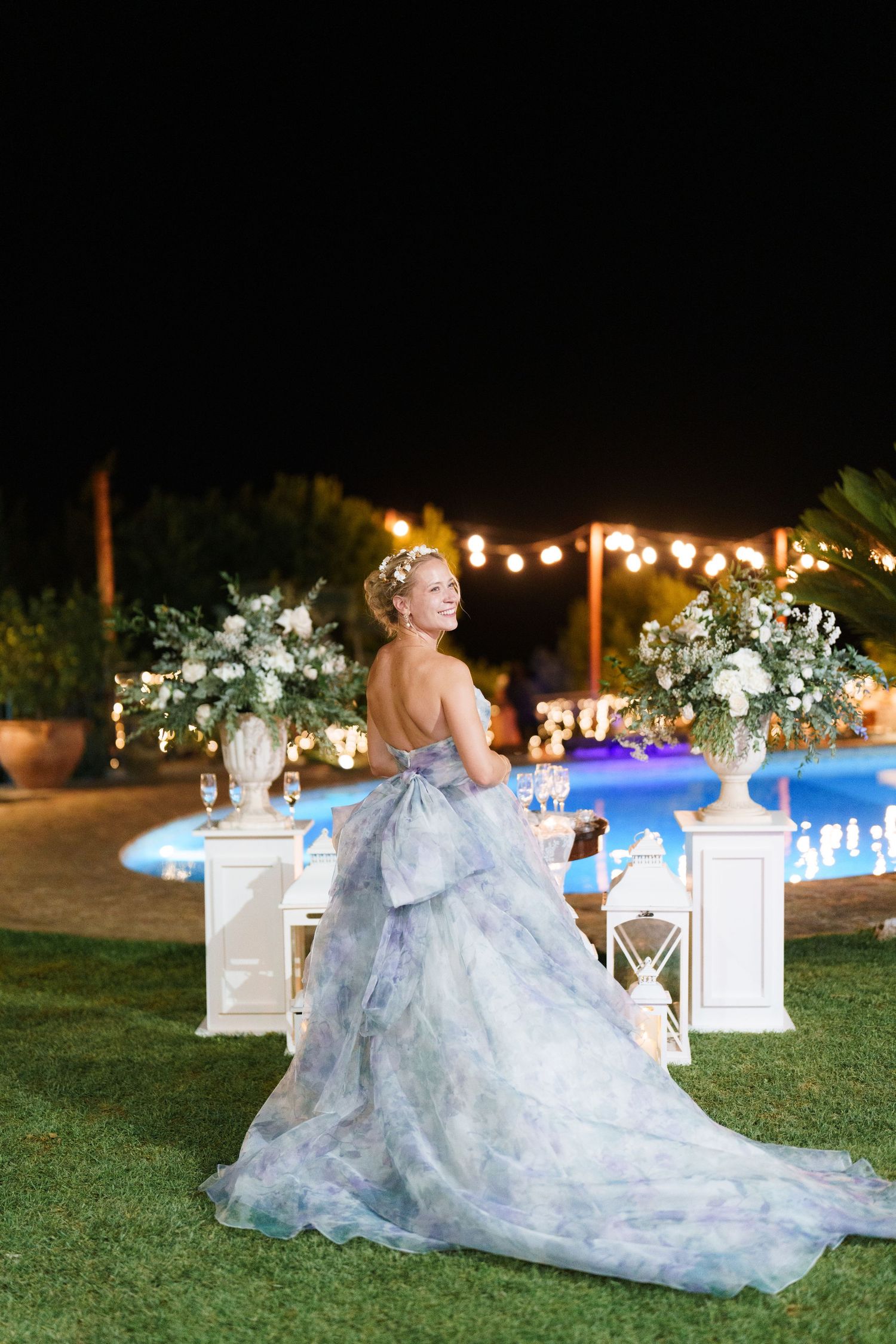 Bride in blue floral gown poses by illuminated pool at night wedding reception with white floral arrangements.