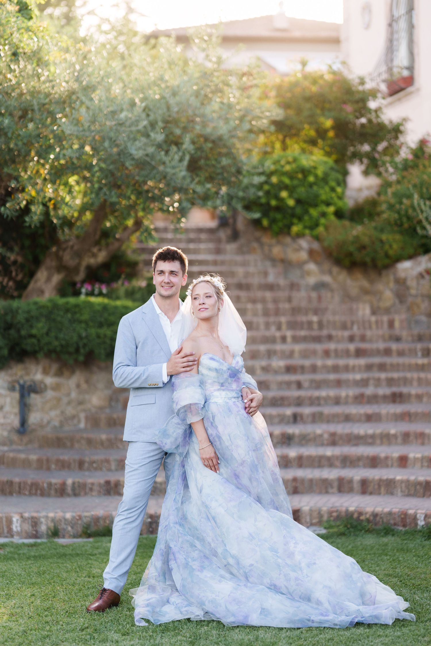 Bride in blue floral gown and groom in gray suit pose on garden steps at outdoor wedding ceremony.
