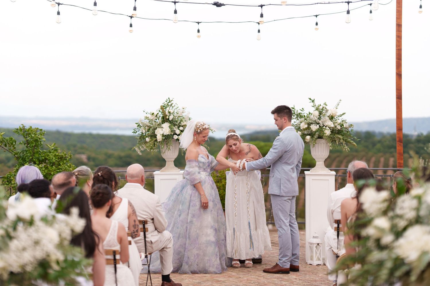 Outdoor wedding ceremony with bride in blue gown, groom in gray suit, and officiant at floral altar with string lights.