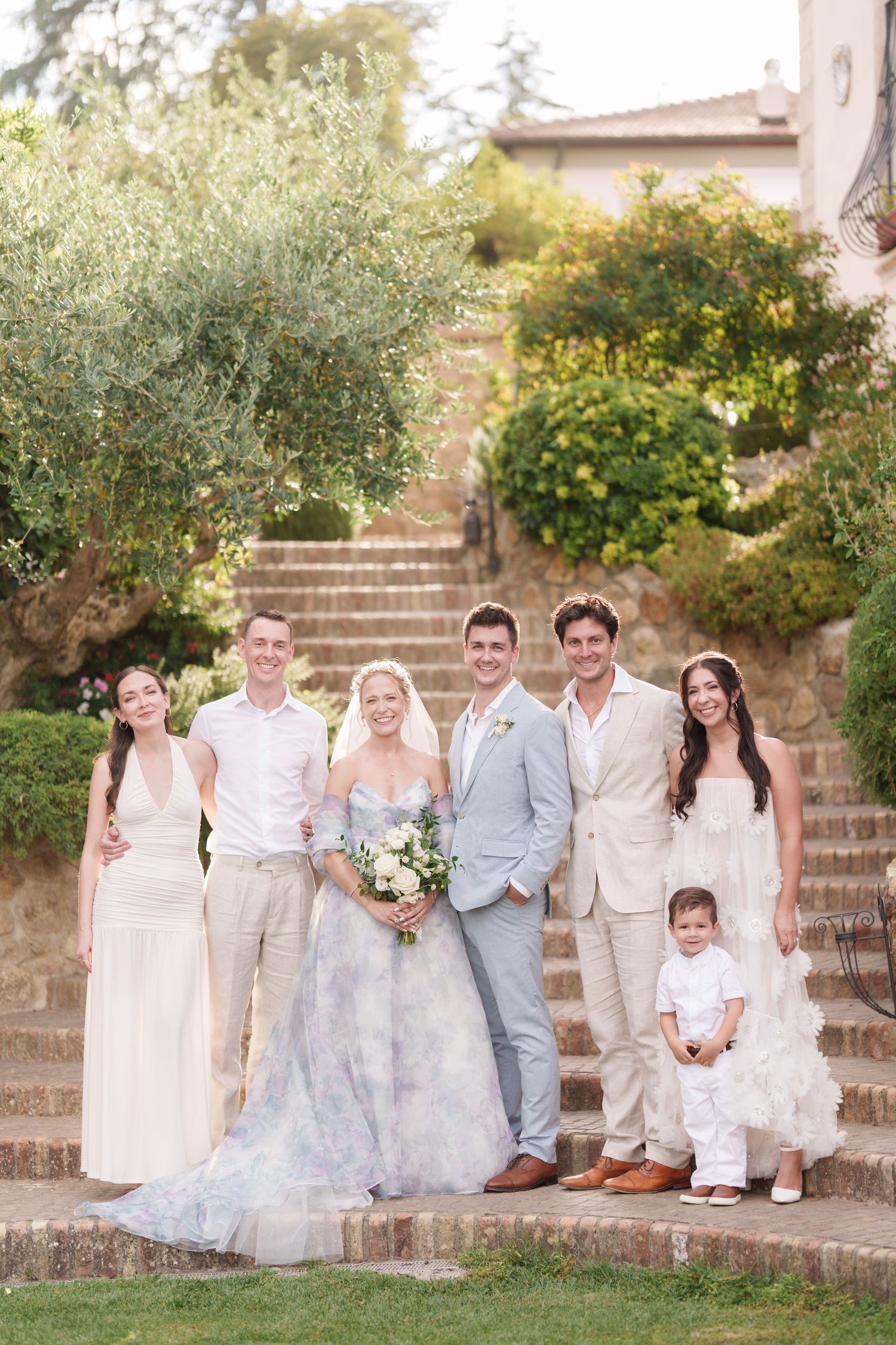 Wedding party posing on stone steps at an outdoor venue, bride in floral gown holding bouquet with guests in elegant attire.
