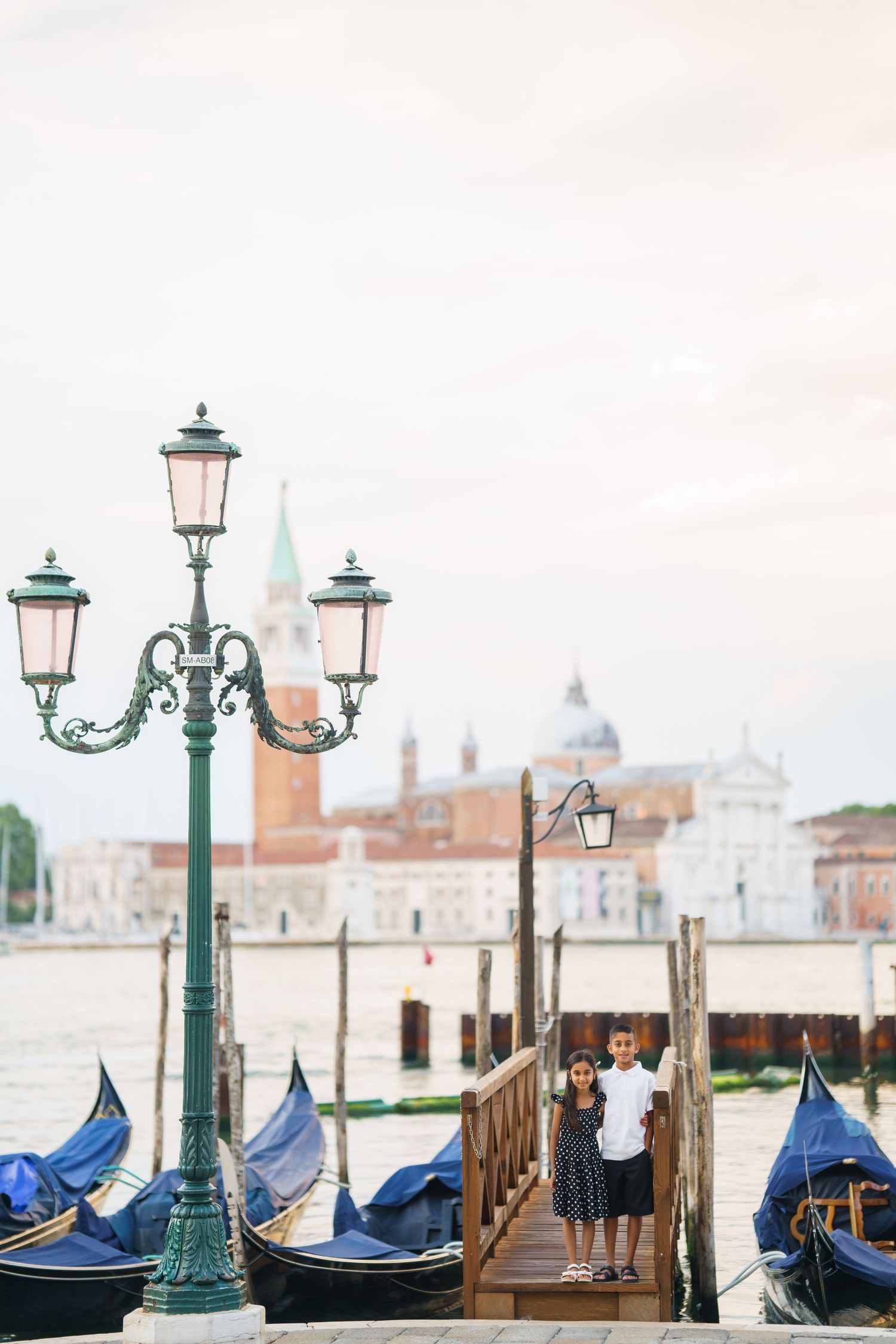 Two children stand on a wooden dock in Venice, Italy, with gondolas and San Giorgio Maggiore church in background.