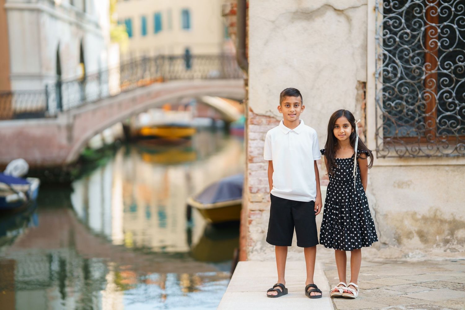 Two children posing by a Venice canal with historic bridges and colorful boats in the background.