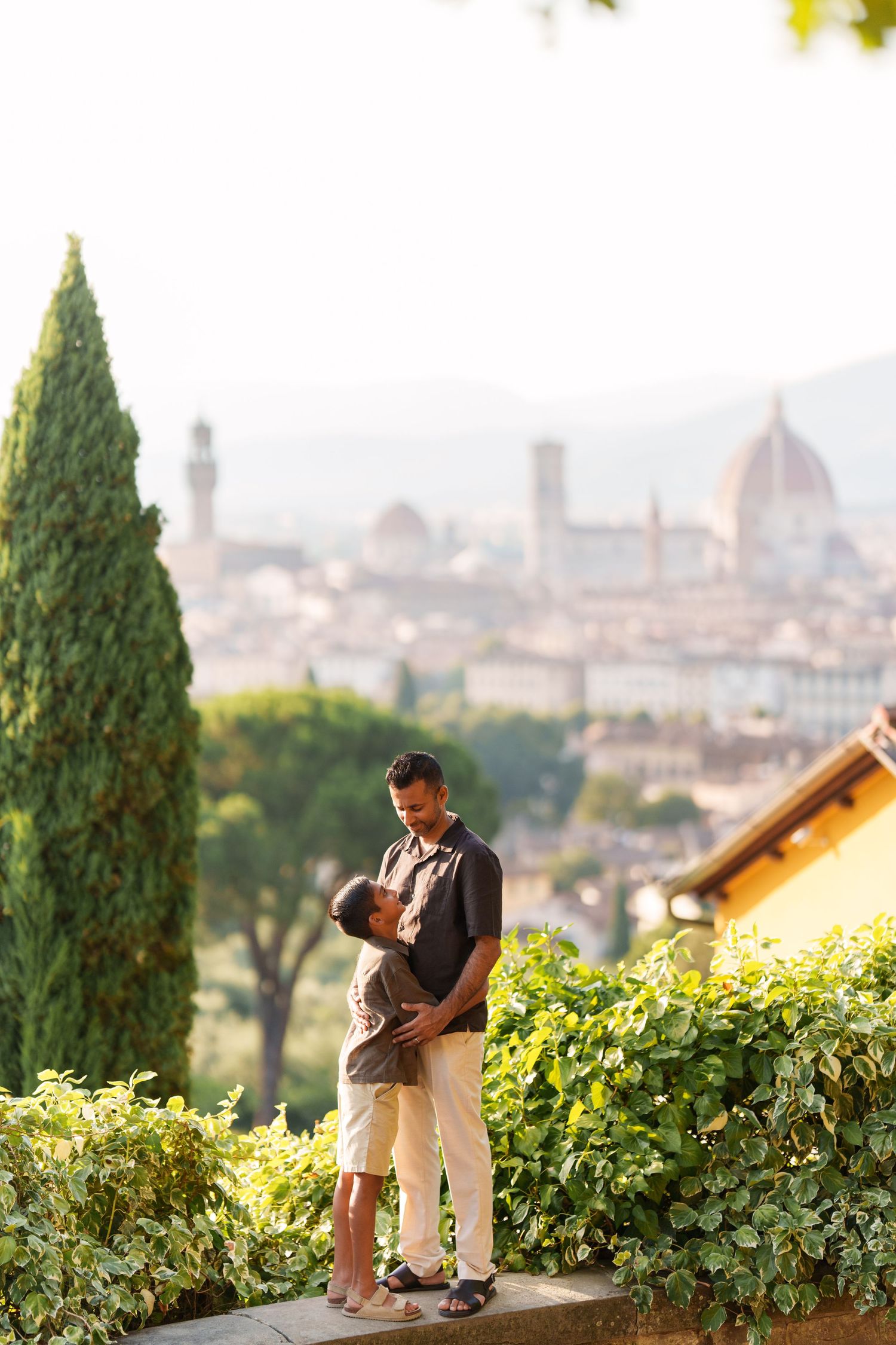 Father and child embrace on a terrace overlooking Florence skyline with cathedral dome and cypress trees.