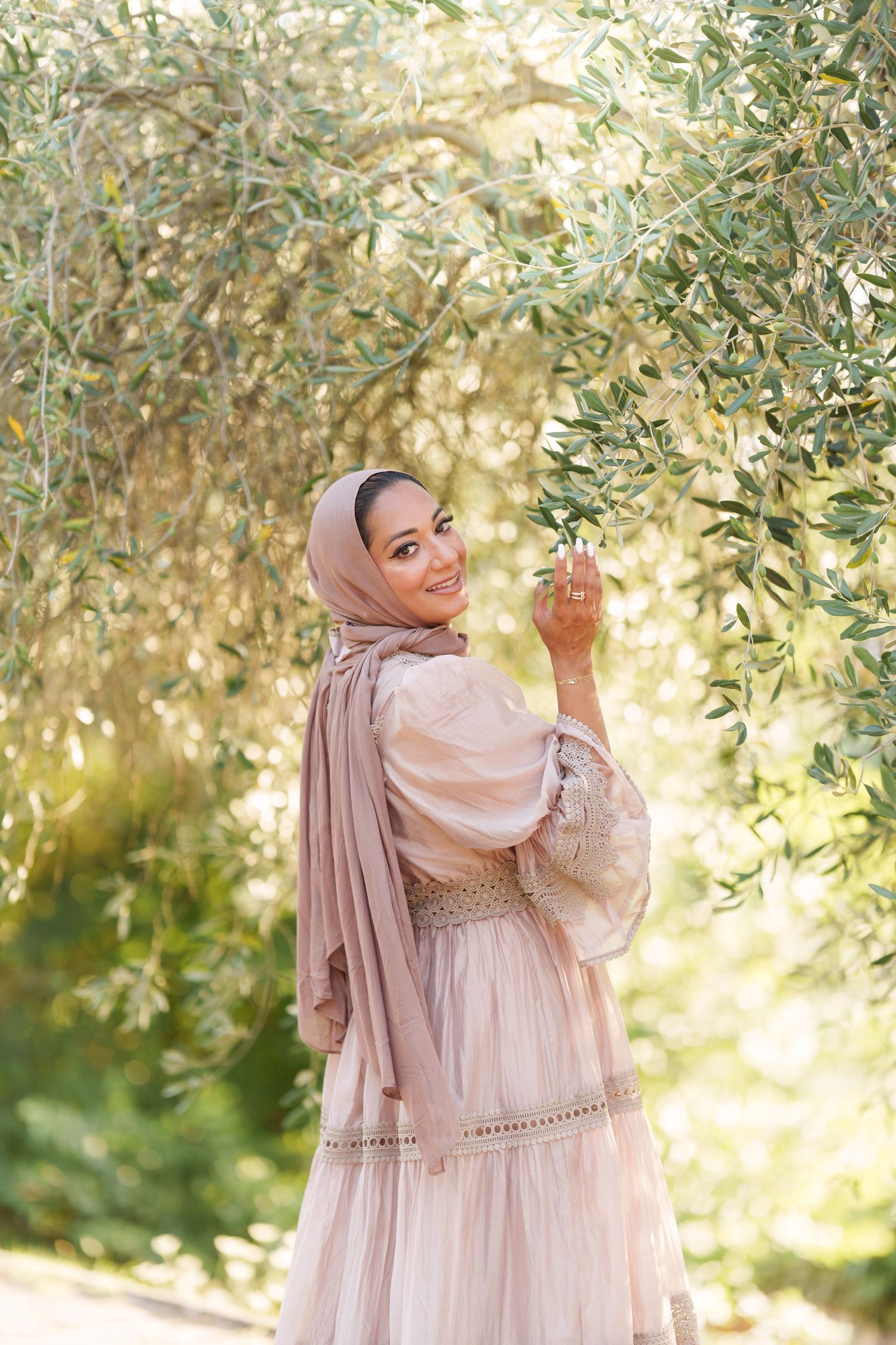 Smiling woman in pink hijab and dress touching olive tree branches in sunlit garden.