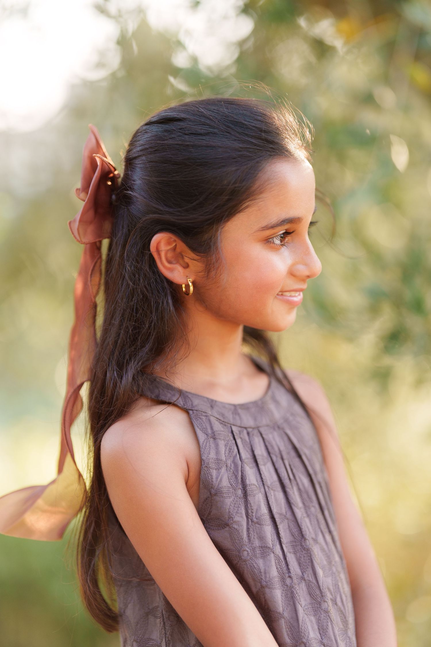 Young girl with long dark hair and ribbon, wearing gray dress, smiling outdoors in natural sunlight.