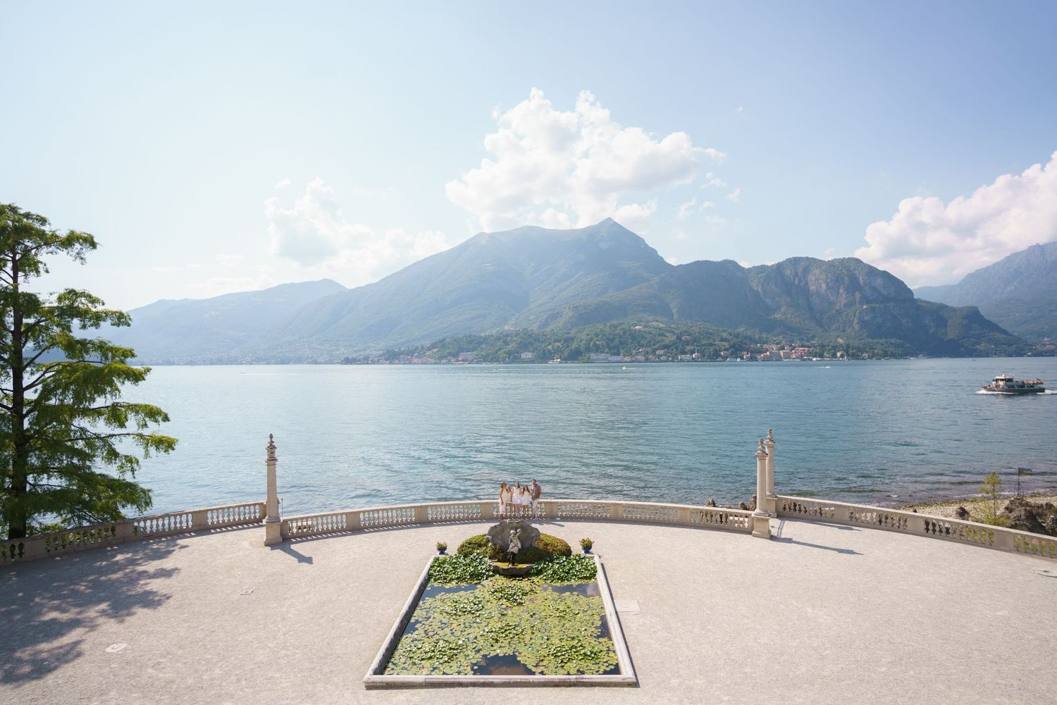 Elegant lakeside terrace with fountain, overlooking serene alpine lake and mountain backdrop under a partly cloudy sky.
