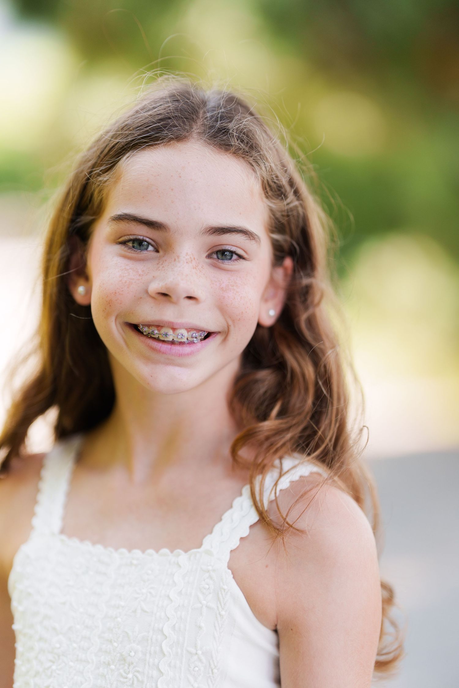 Smiling young girl with braces, freckles, and brown hair wearing a white dress outdoors.
