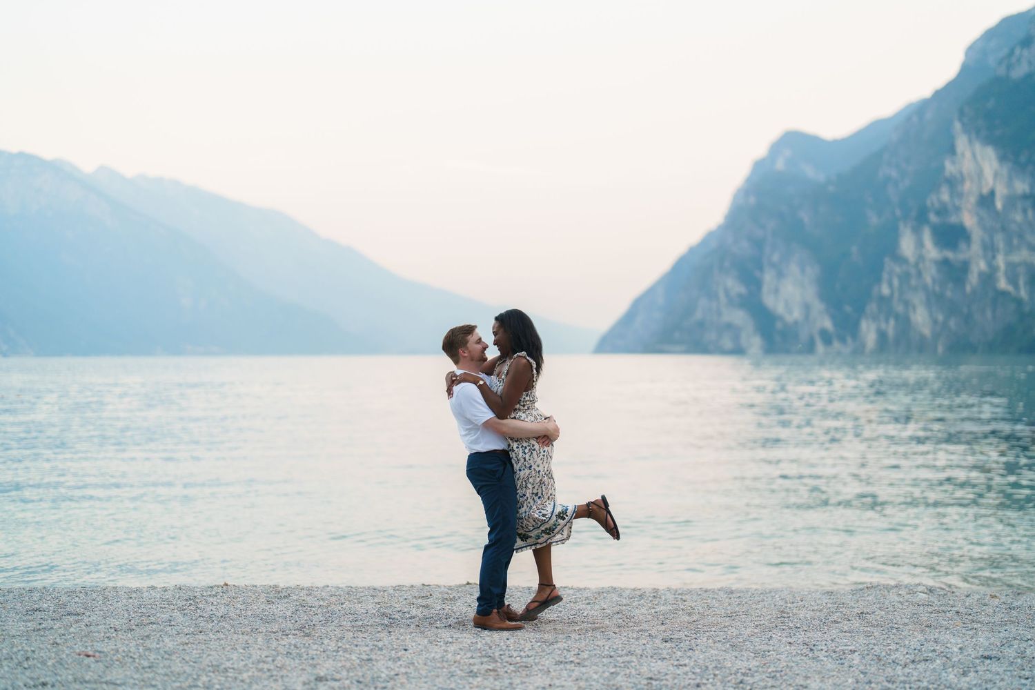 Romantic couple kissing on Lake Garda beach with breathtaking alpine mountain backdrop.