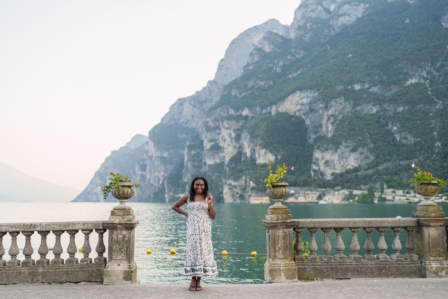 Elegant woman in white floral dress posing on stone balustrade overlooking beautiful Lake Garda, Italy.