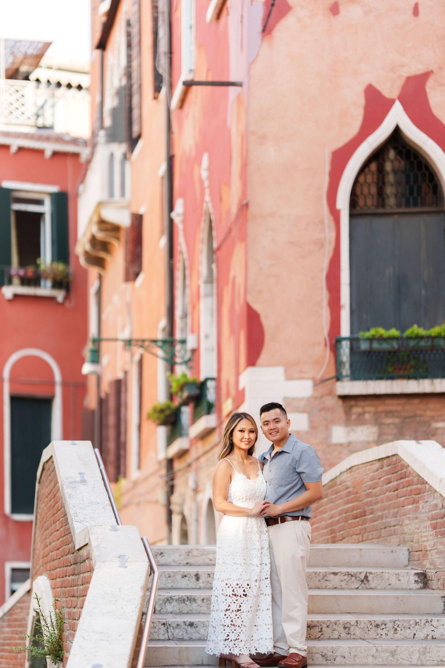 Couple embracing on stone steps in Venice, Italy, with colorful pink and red historic buildings in the background.