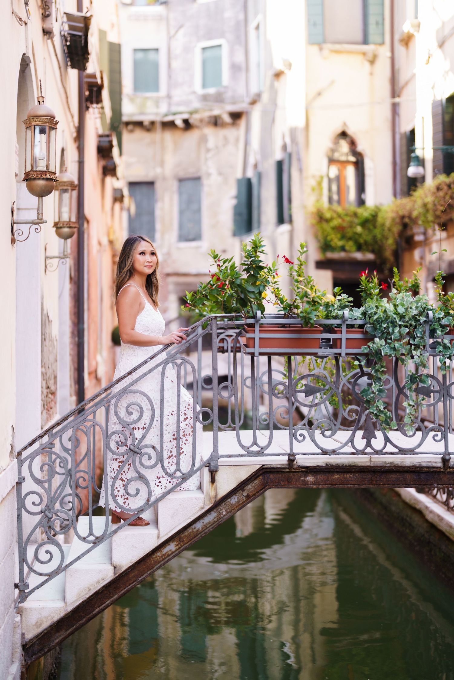 Woman in white dress standing on ornate iron bridge over Venice canal surrounded by colorful flowers and historic buildings.