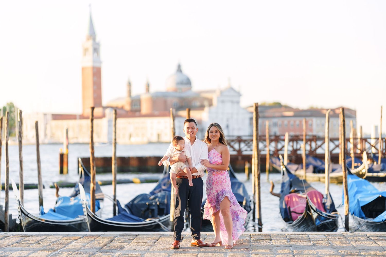 Family of three posing by gondolas on Venice waterfront with San Giorgio Maggiore church in background.