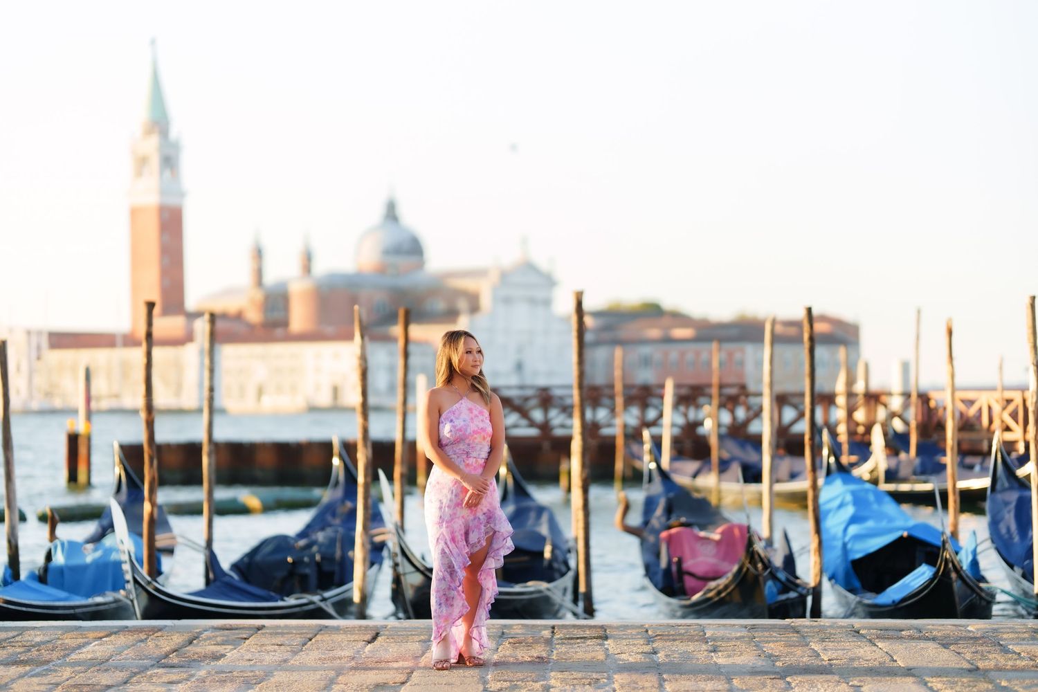 Woman in pink dress posing by Venice gondolas with San Giorgio Maggiore church in background at sunset.