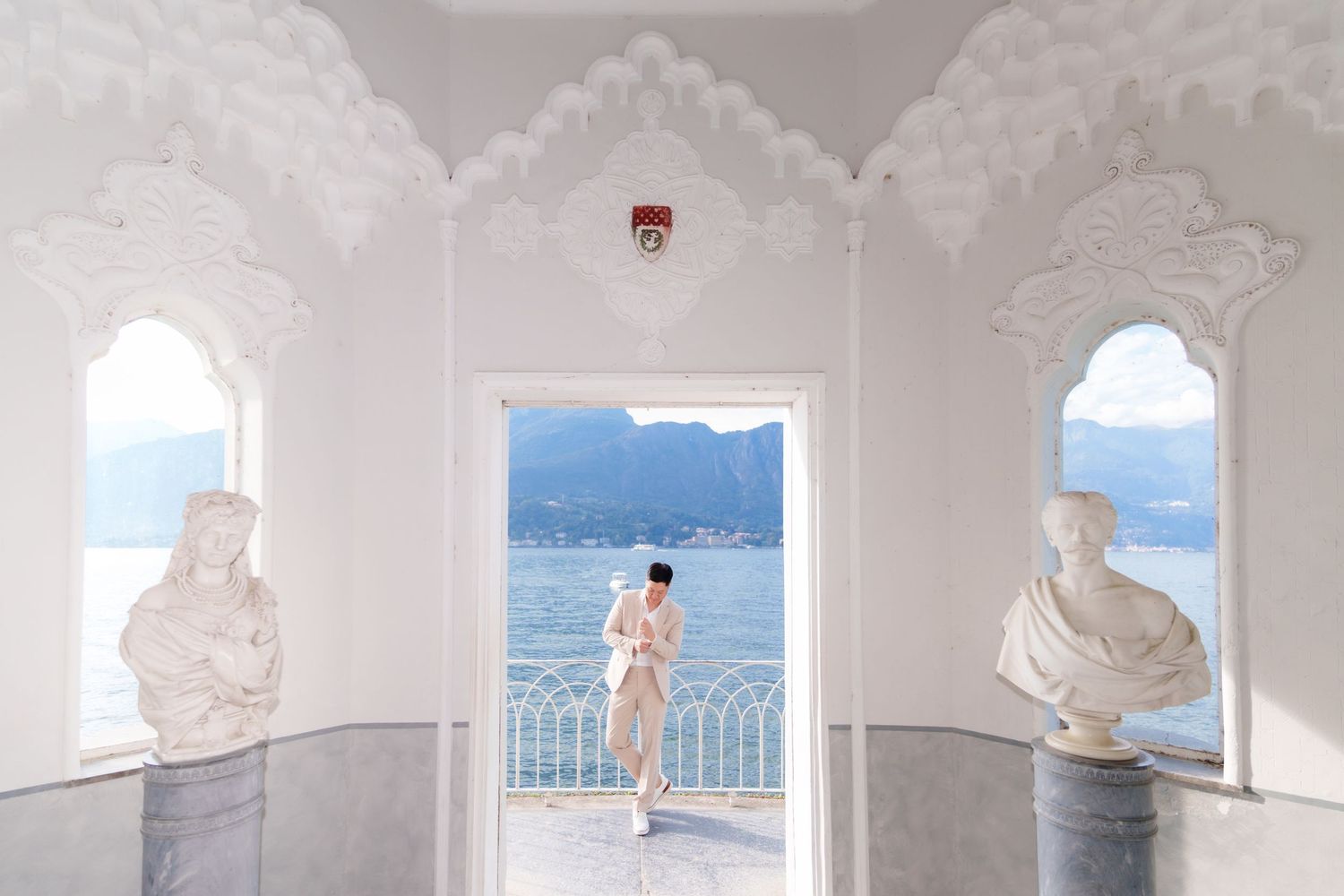 Figure stands in ornate white archway overlooking a serene lake with mountains in the background.