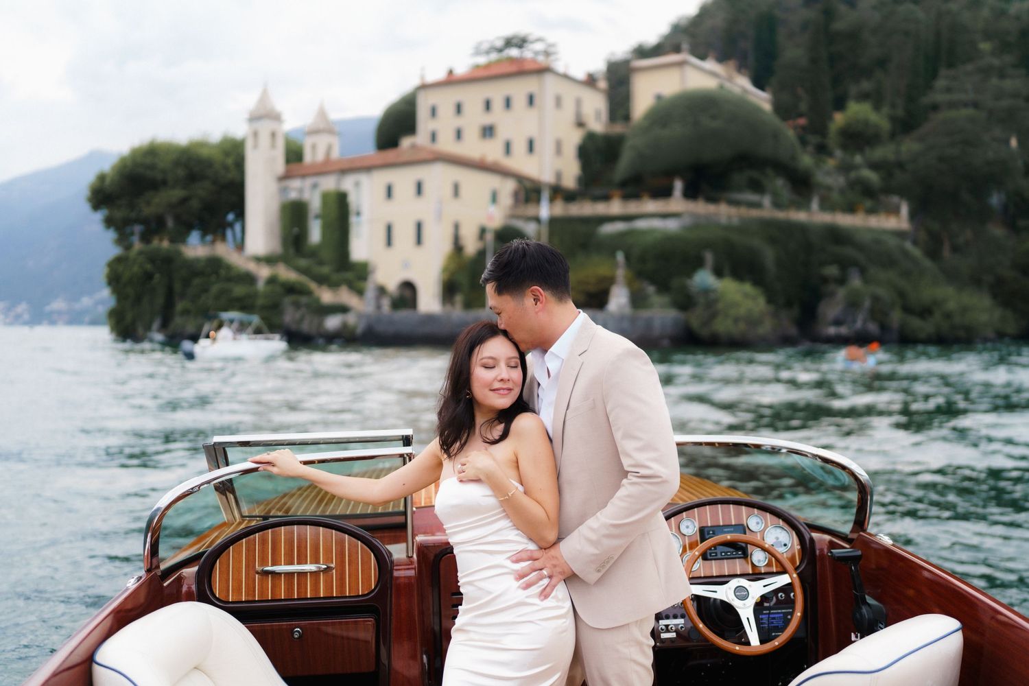 A romantic couple poses together in a wooden boat on Lake Como in Italy with scenic views of historic villas in the background.