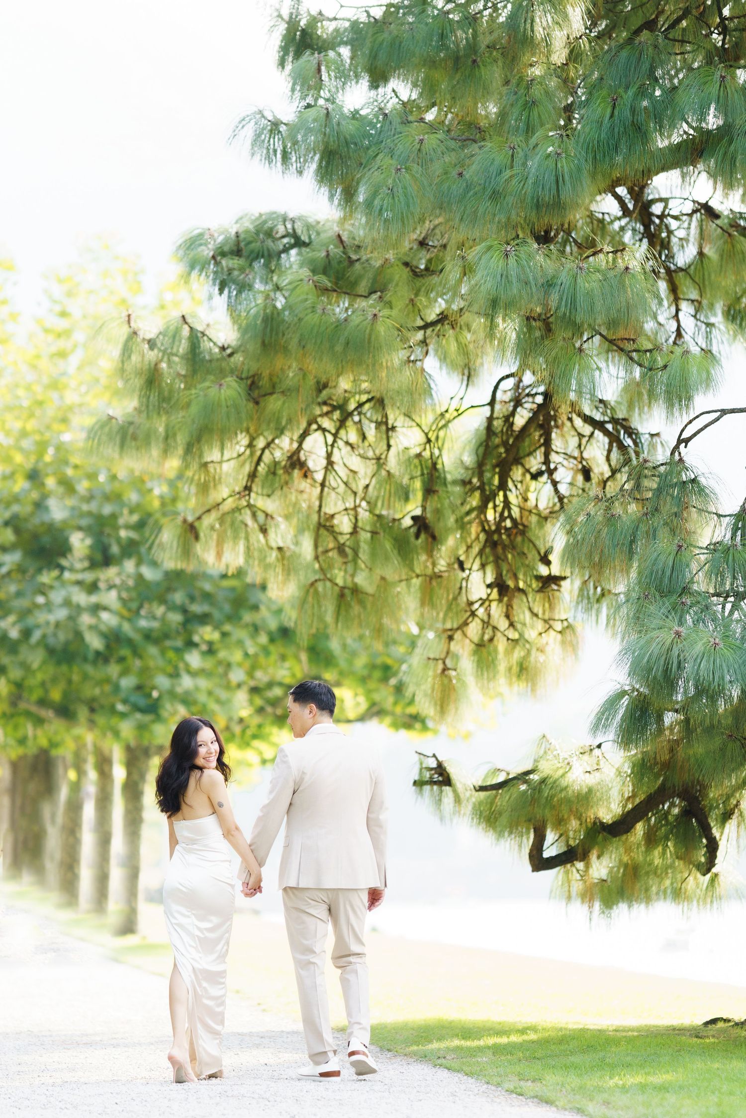 Couple in white outfits walking together under green trees in a romantic outdoor setting.