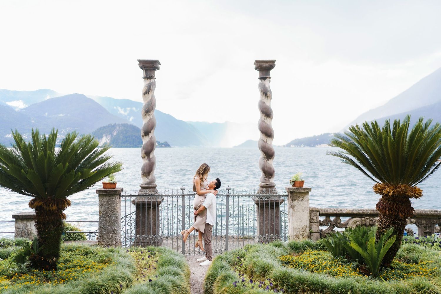 A romantic couple shares a tender moment on a scenic terrace overlooking Lake Como in Italy with mountains in the background.