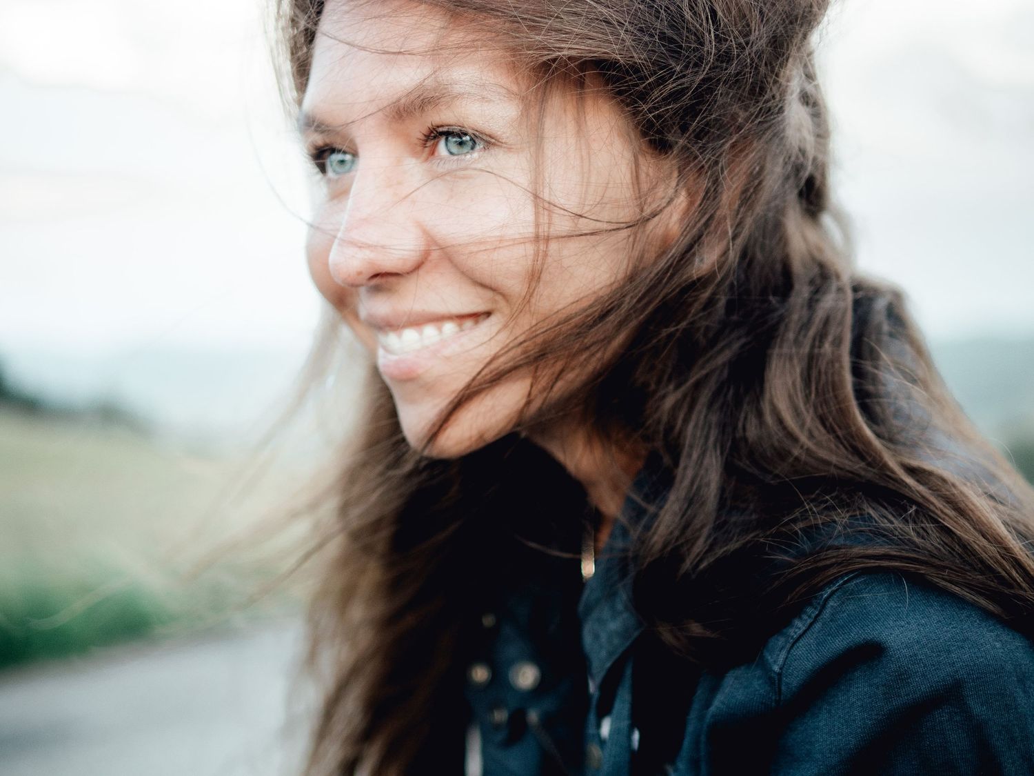 A person with long dark hair smiles while looking away from the camera in a black jacket outdoors.