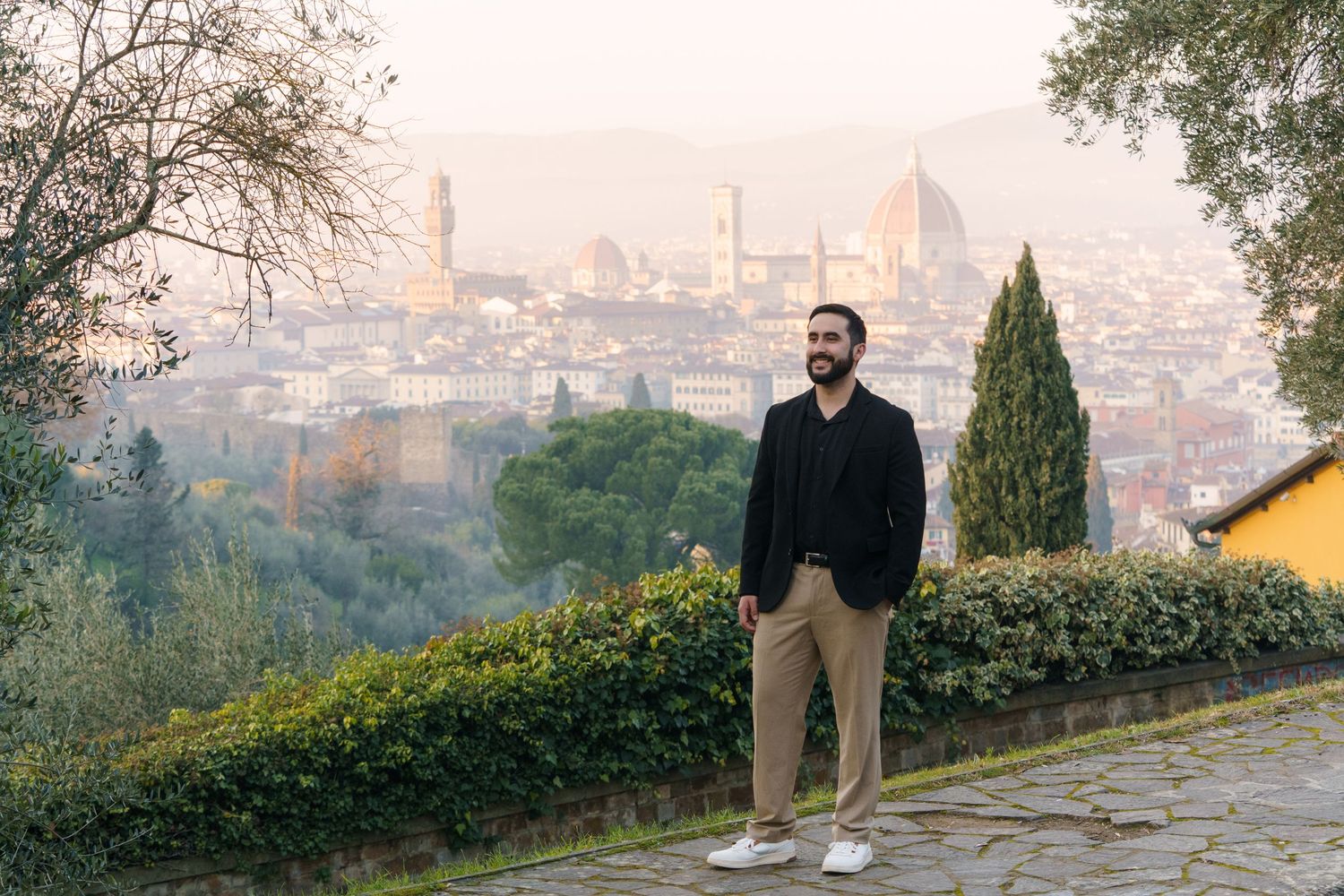 A person walks along a scenic hilltop path overlooking a city skyline at sunset with cypress trees in the foreground.