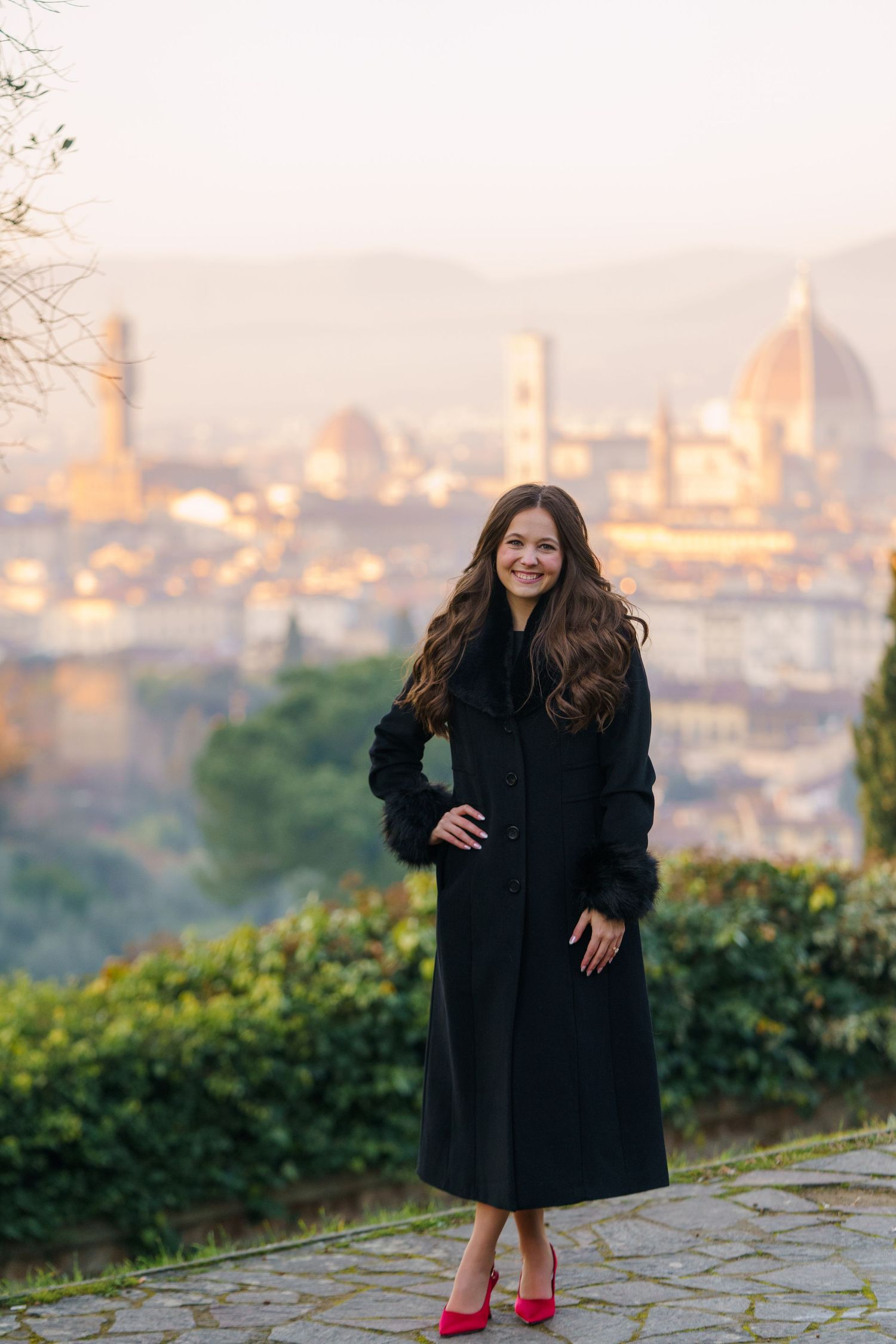 A person in a black coat and red shoes poses on a scenic overlook above Florence, Italy with the Duomo visible in the distance.