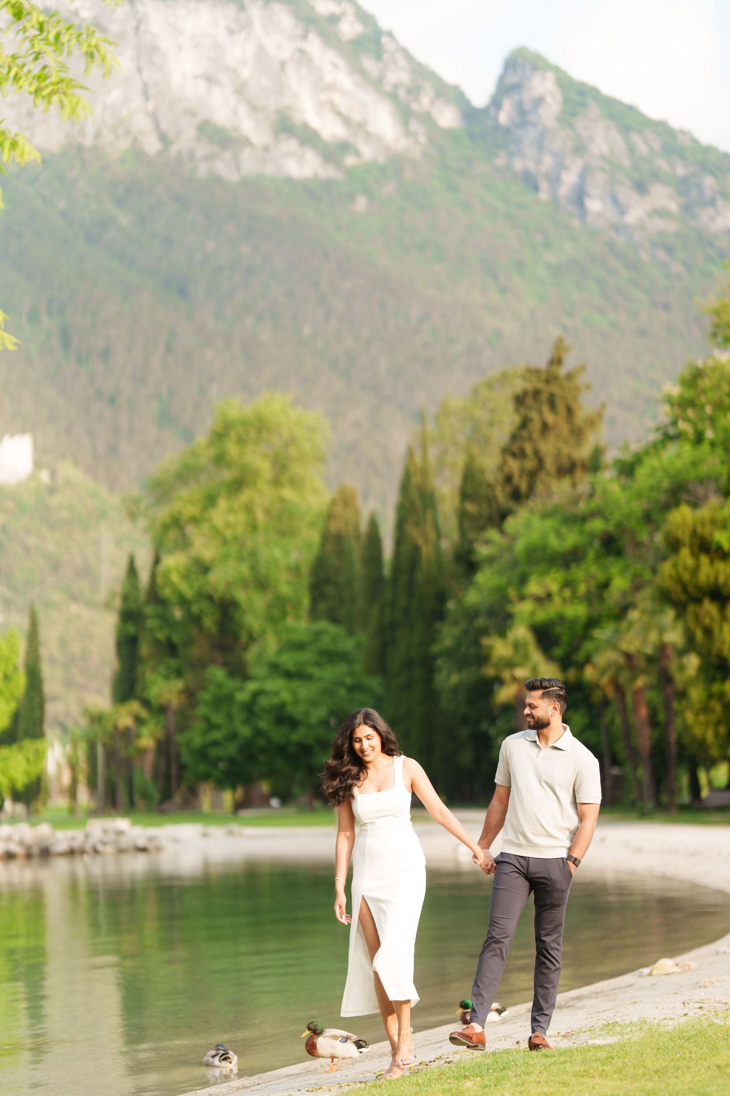 Couple walking along scenic lakeside path with mountains and lush greenery in the background.