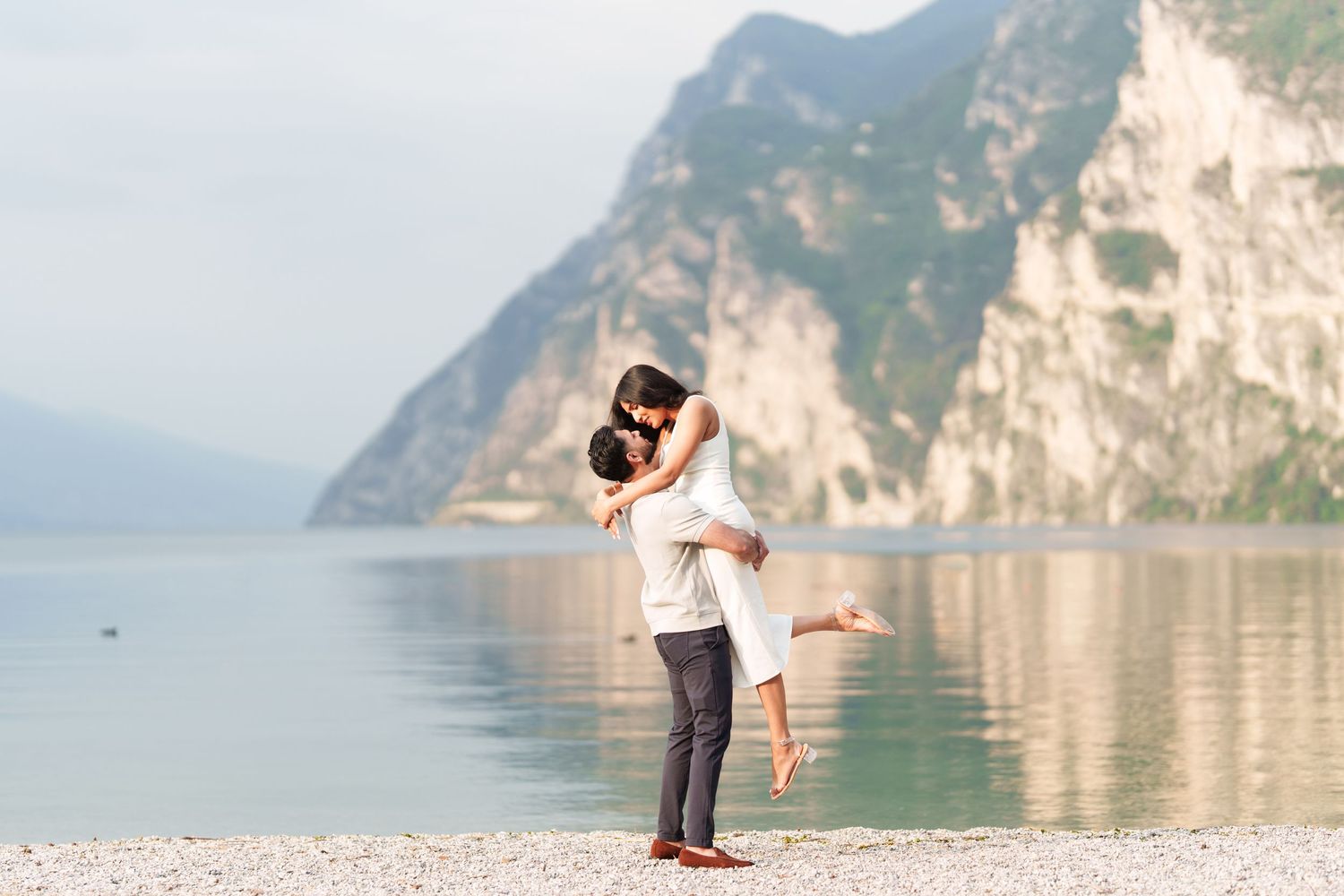 A romantic couple embraces against the backdrop of Lake Garda in Italy with dramatic mountain cliffs and serene waters.