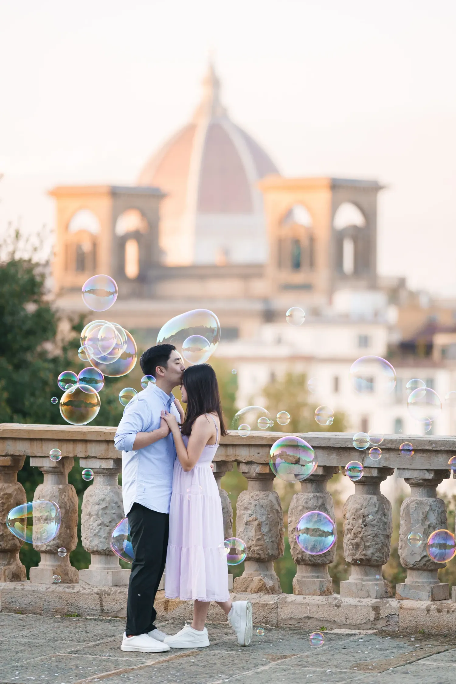 Two children play with bubbles near a stone balustrade at sunset with a castle-like building in the background.