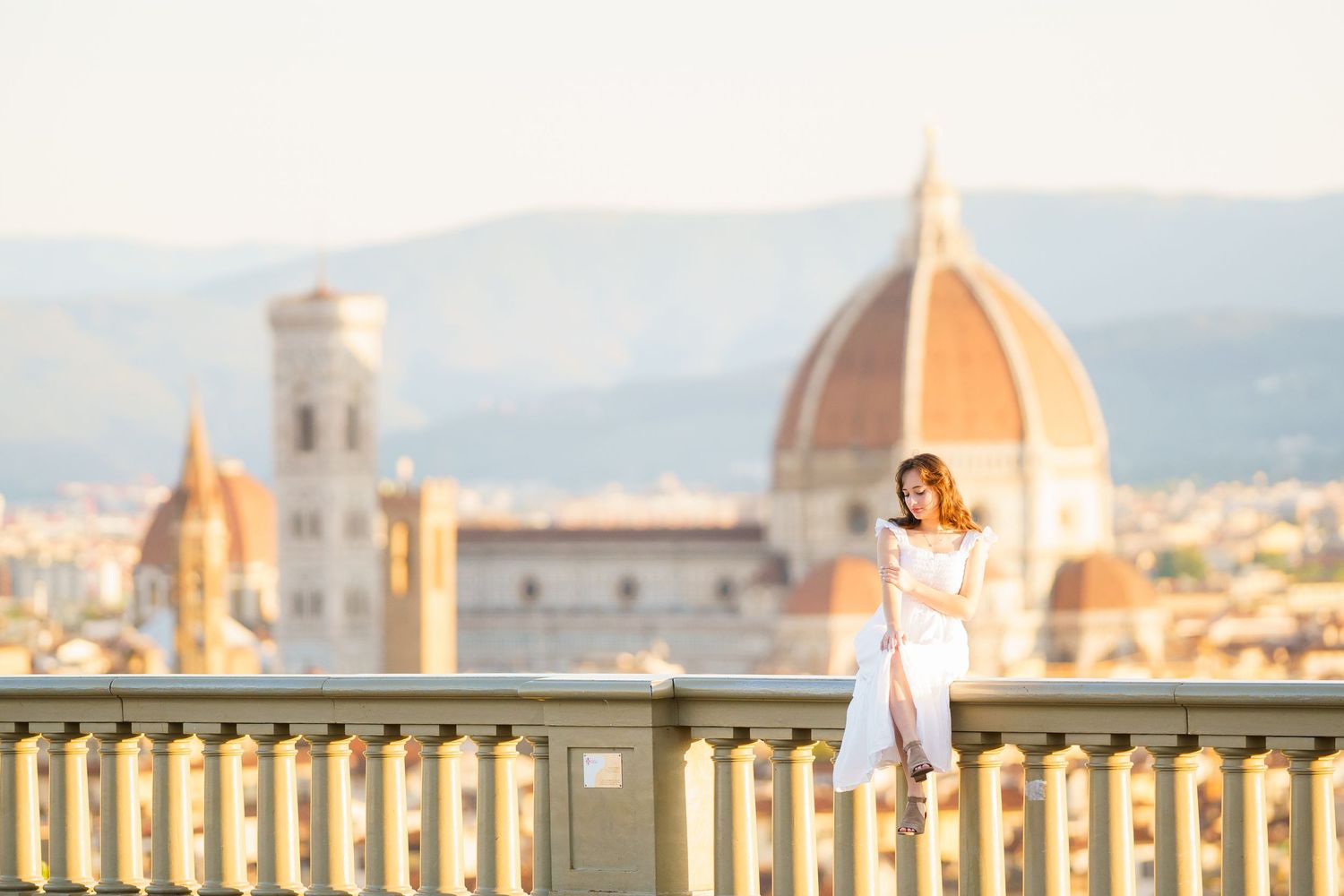 A person in a white dress poses on a balcony overlooking Florence's iconic Duomo cathedral during golden hour.