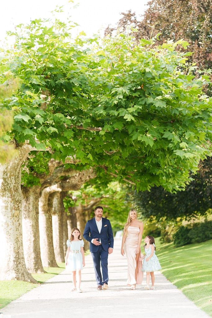 A family walks together down a tree-lined path on a sunny summer day in a peaceful garden setting.