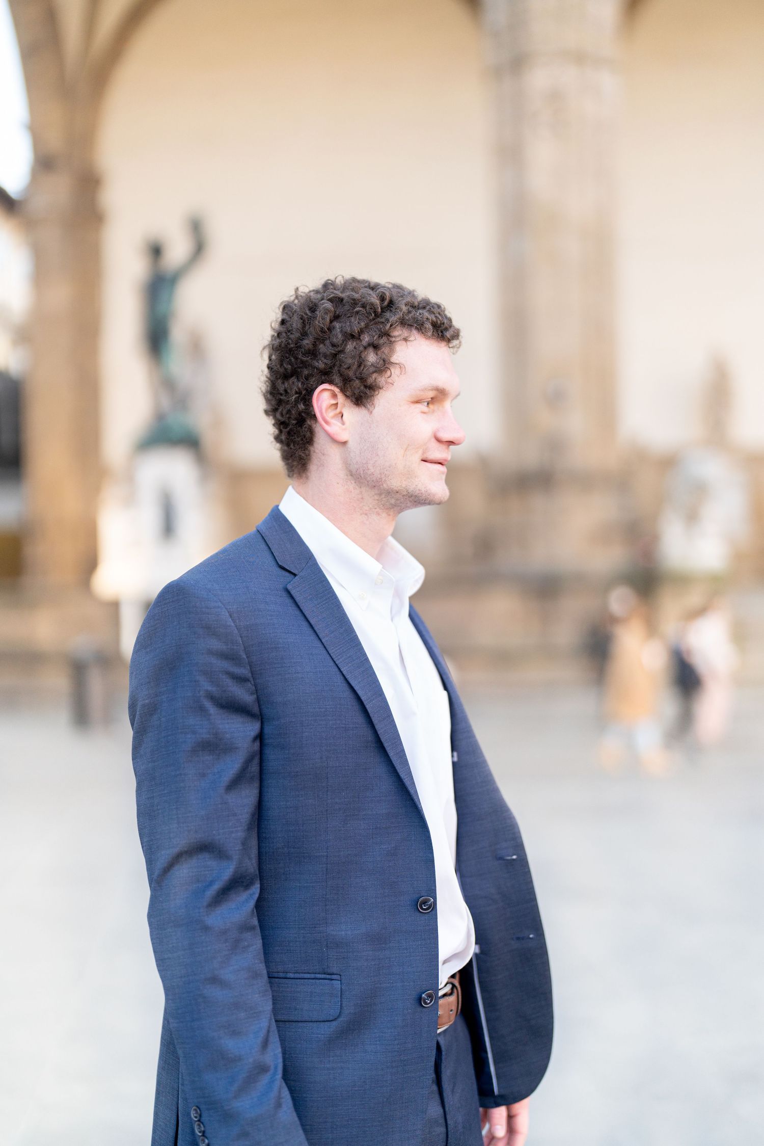 A person in a navy suit and white shirt stands against a light-colored architectural backdrop in a series of photos.