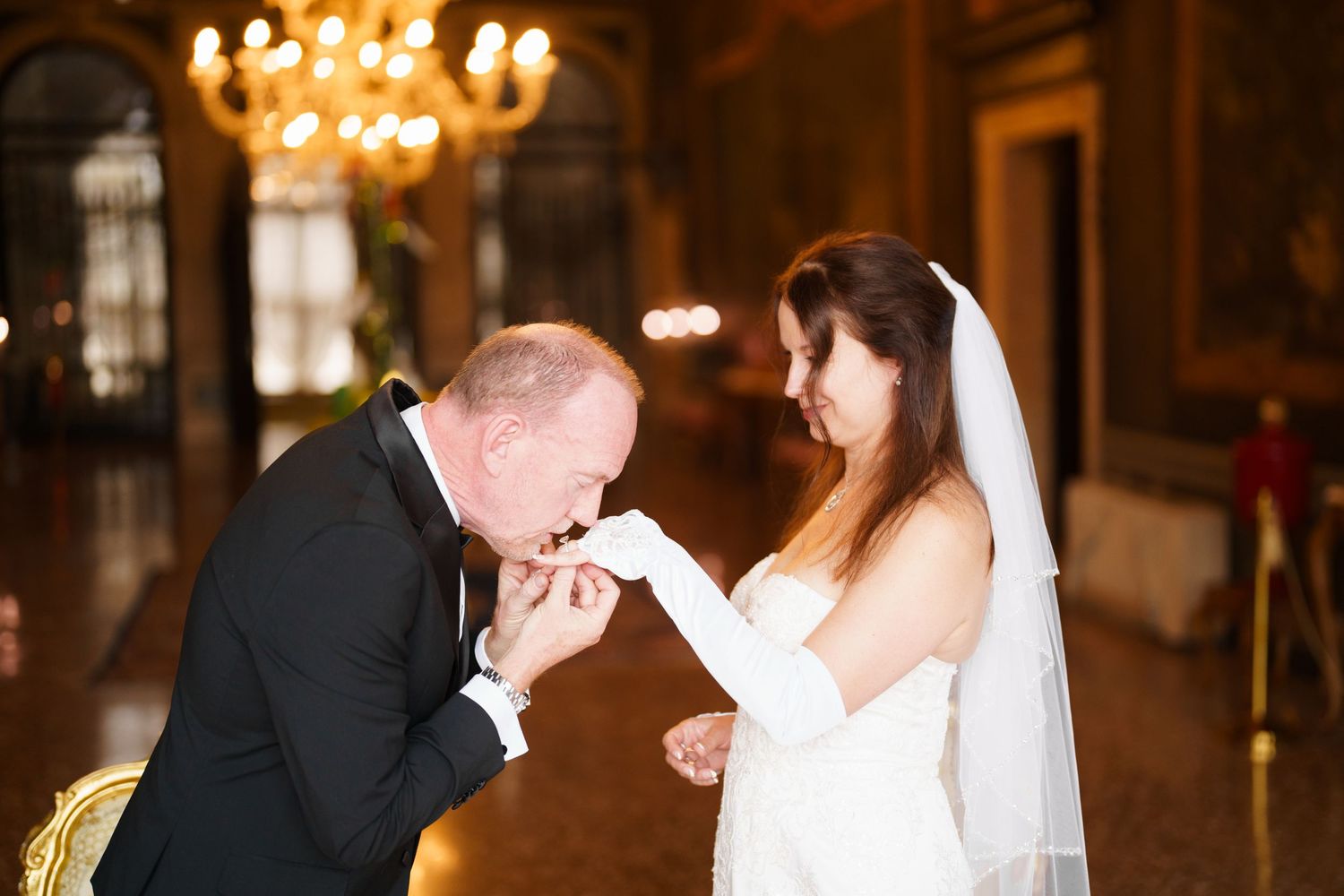A romantic wedding couple shares an intimate moment in a candlelit ballroom with elegant chandeliers overhead.