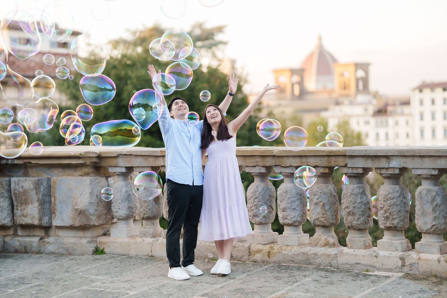 A couple enjoys a romantic moment surrounded by floating soap bubbles on a historic stone balcony at sunset.