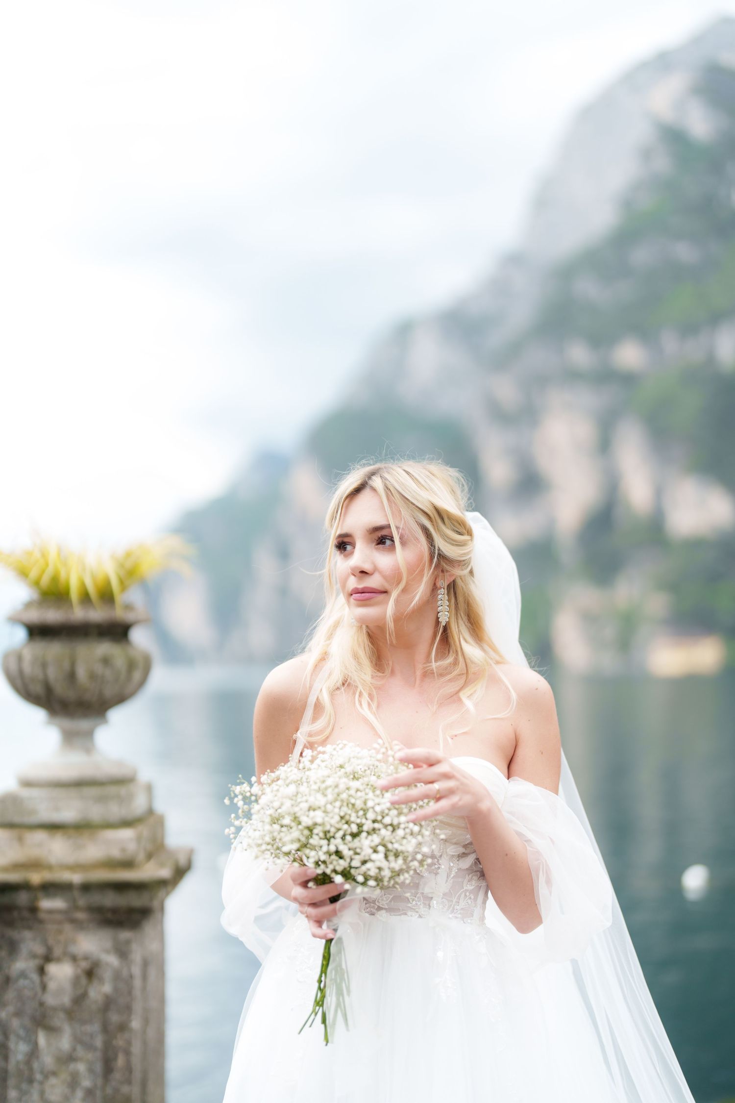 Bride in flowing white dress holds delicate baby's breath bouquet against scenic mountain lake backdrop in Italy.