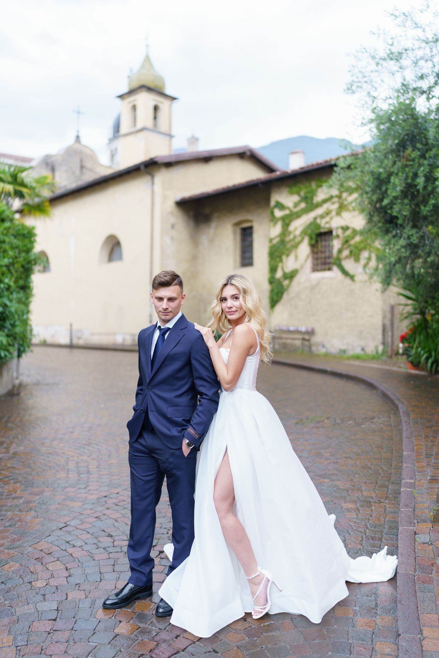 A couple in wedding attire sits together on stone steps outside a church with a tower in the background.