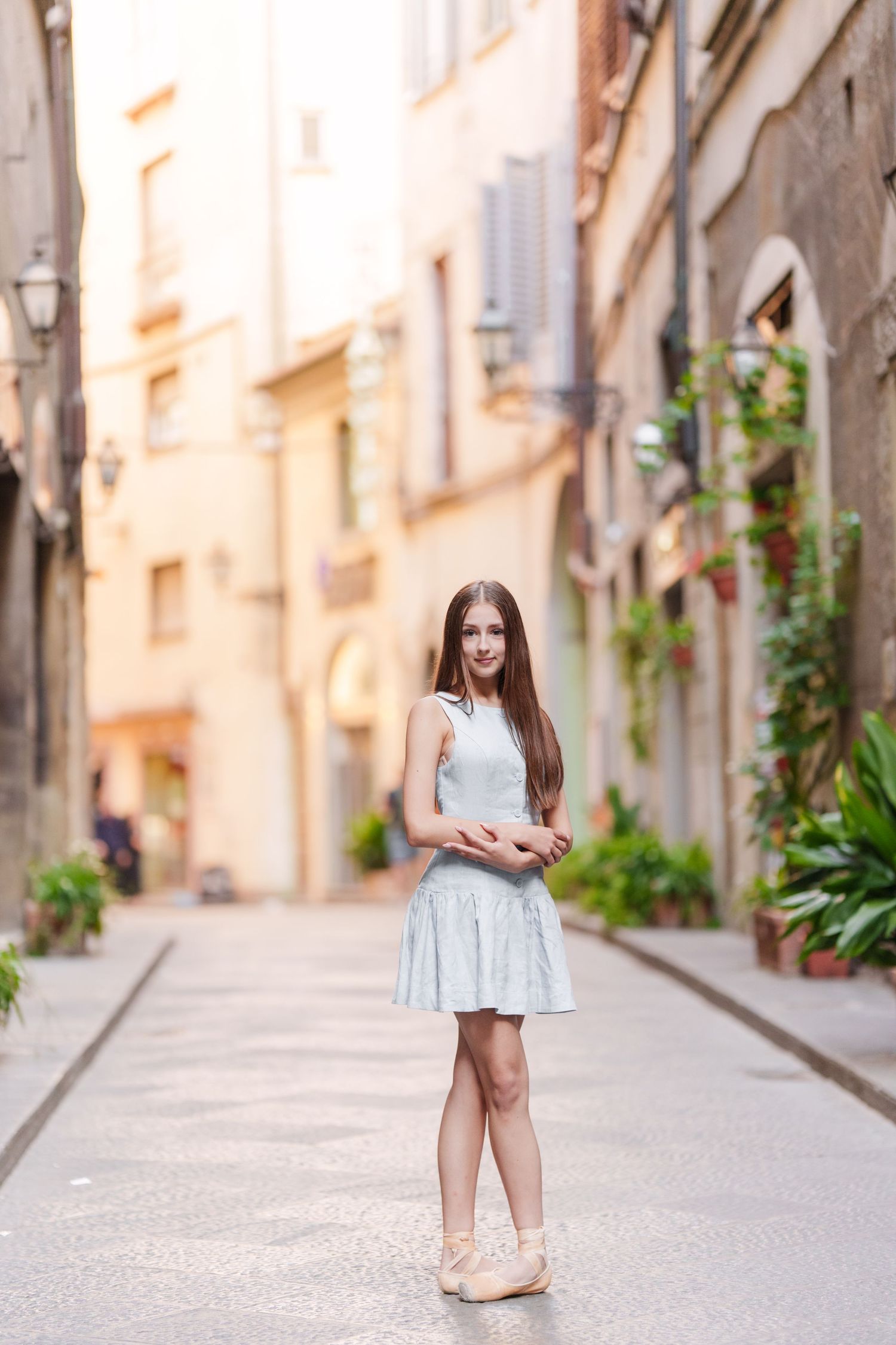 A person in a light blue dress stands in a charming European alleyway with old buildings and plants.