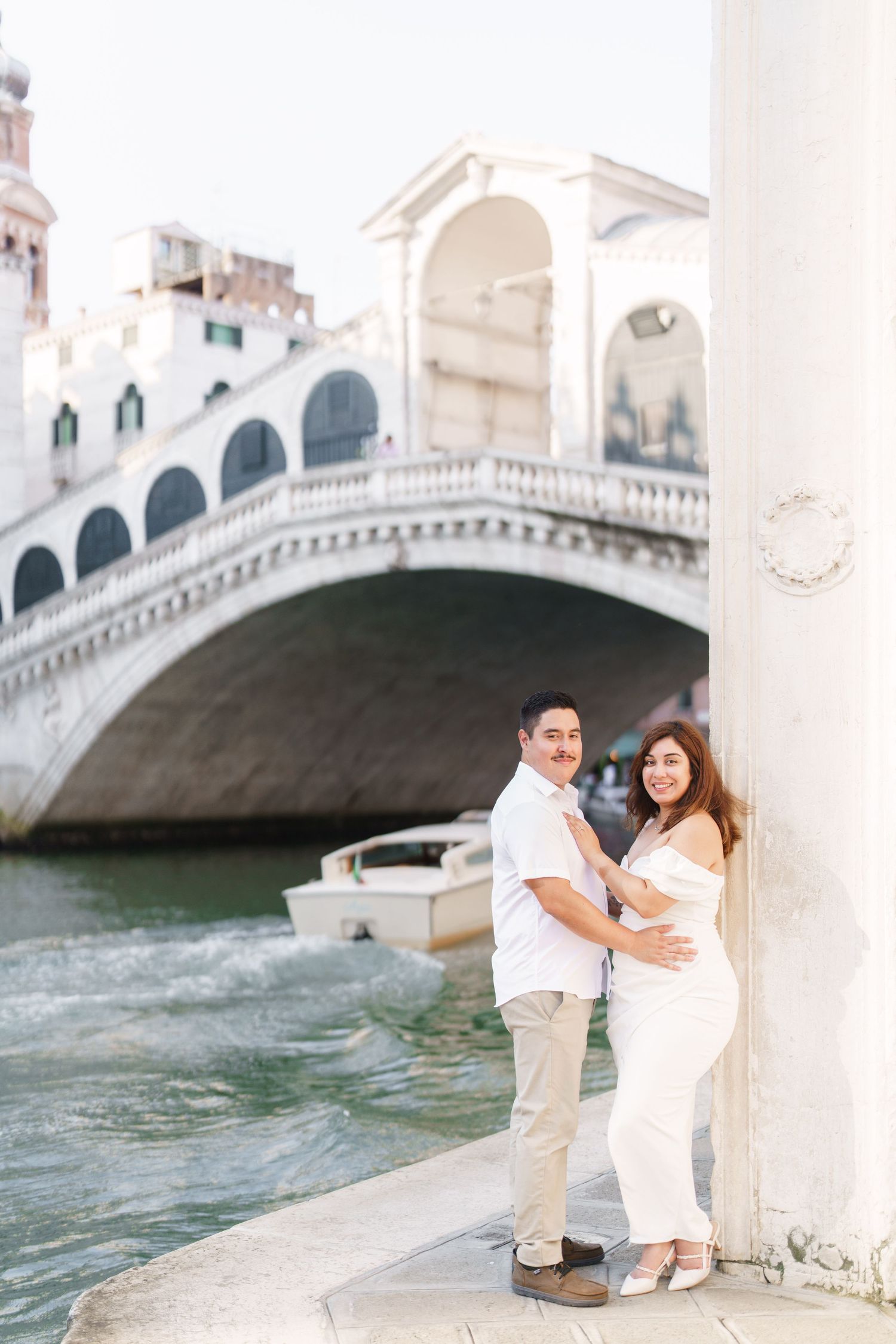 Romantic couple in white outfits posing near the iconic Rialto Bridge in Venice, Italy during a sunny day.