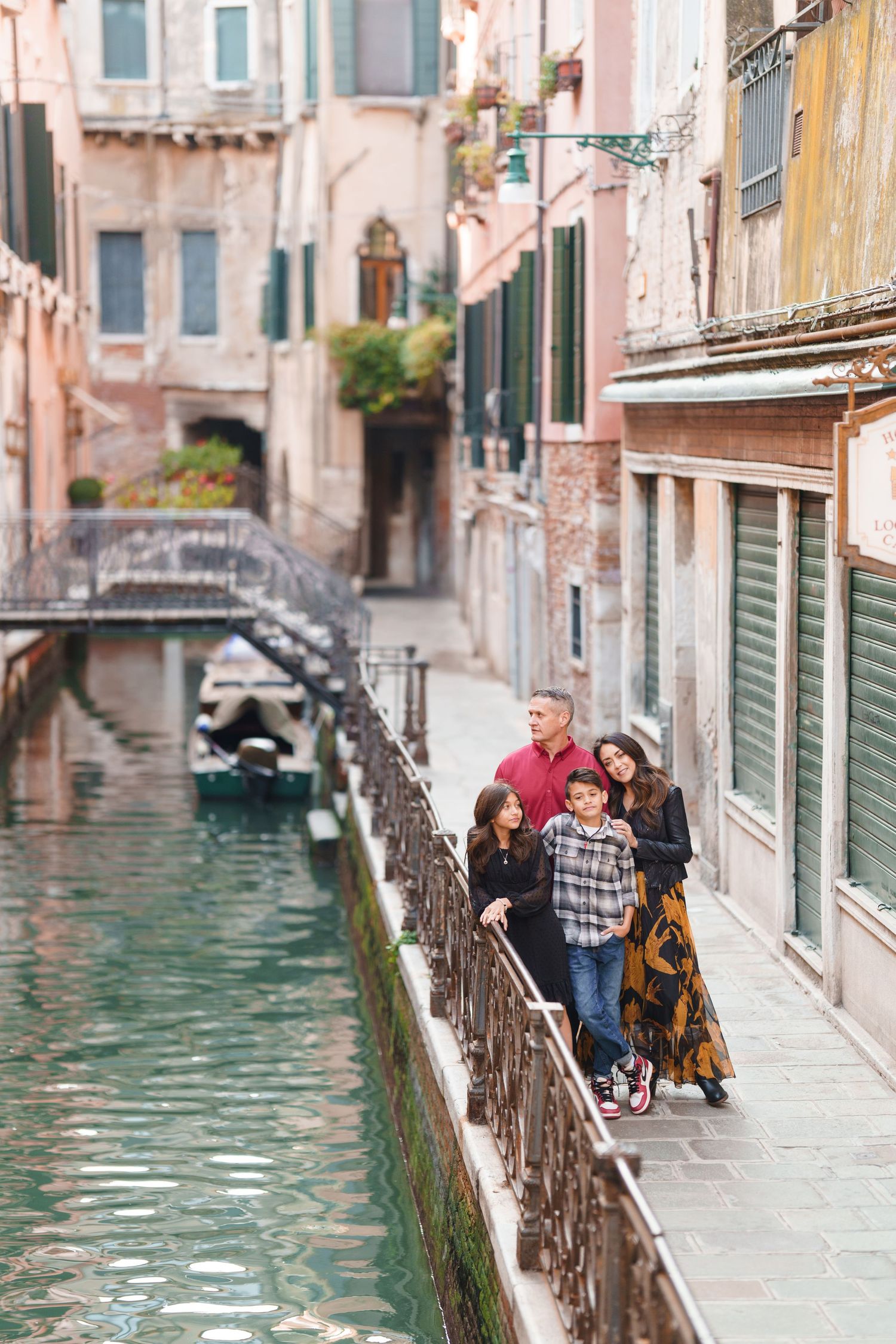 Tourists walk along a narrow stone pathway beside a scenic Venice canal with pink and peach colored buildings.