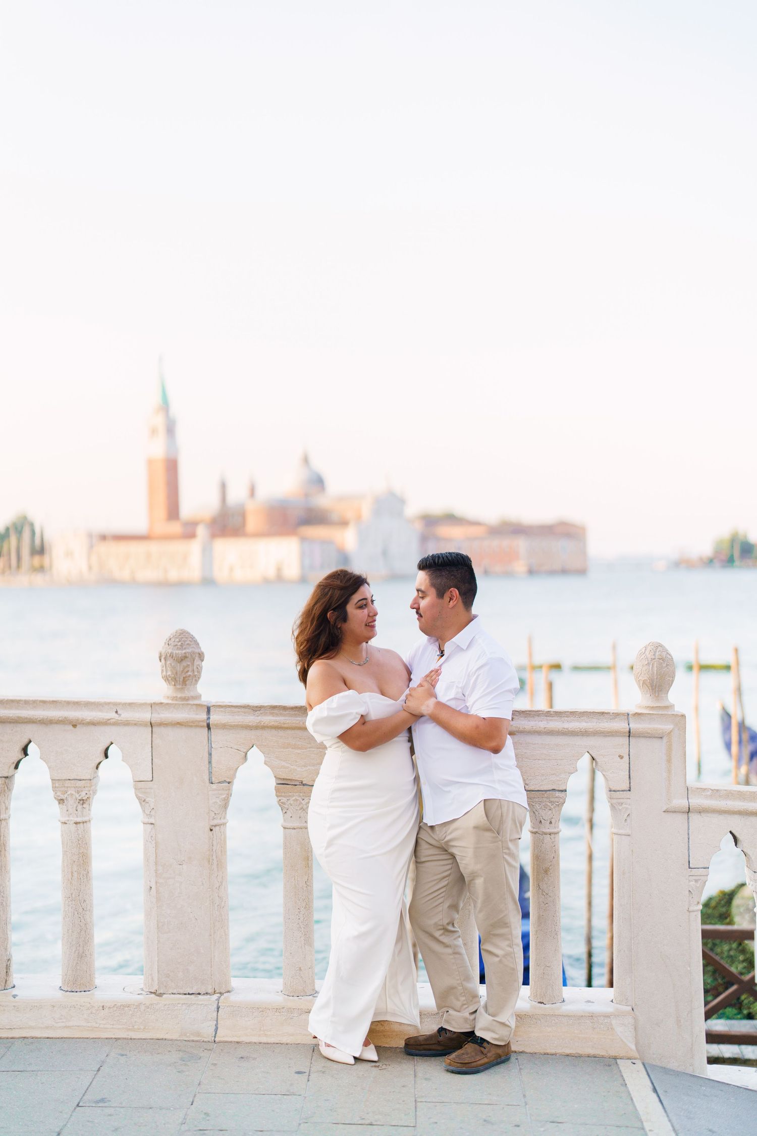 A romantic couple in white outfits shares tender moments along a waterfront balcony with Venice's skyline in the background.