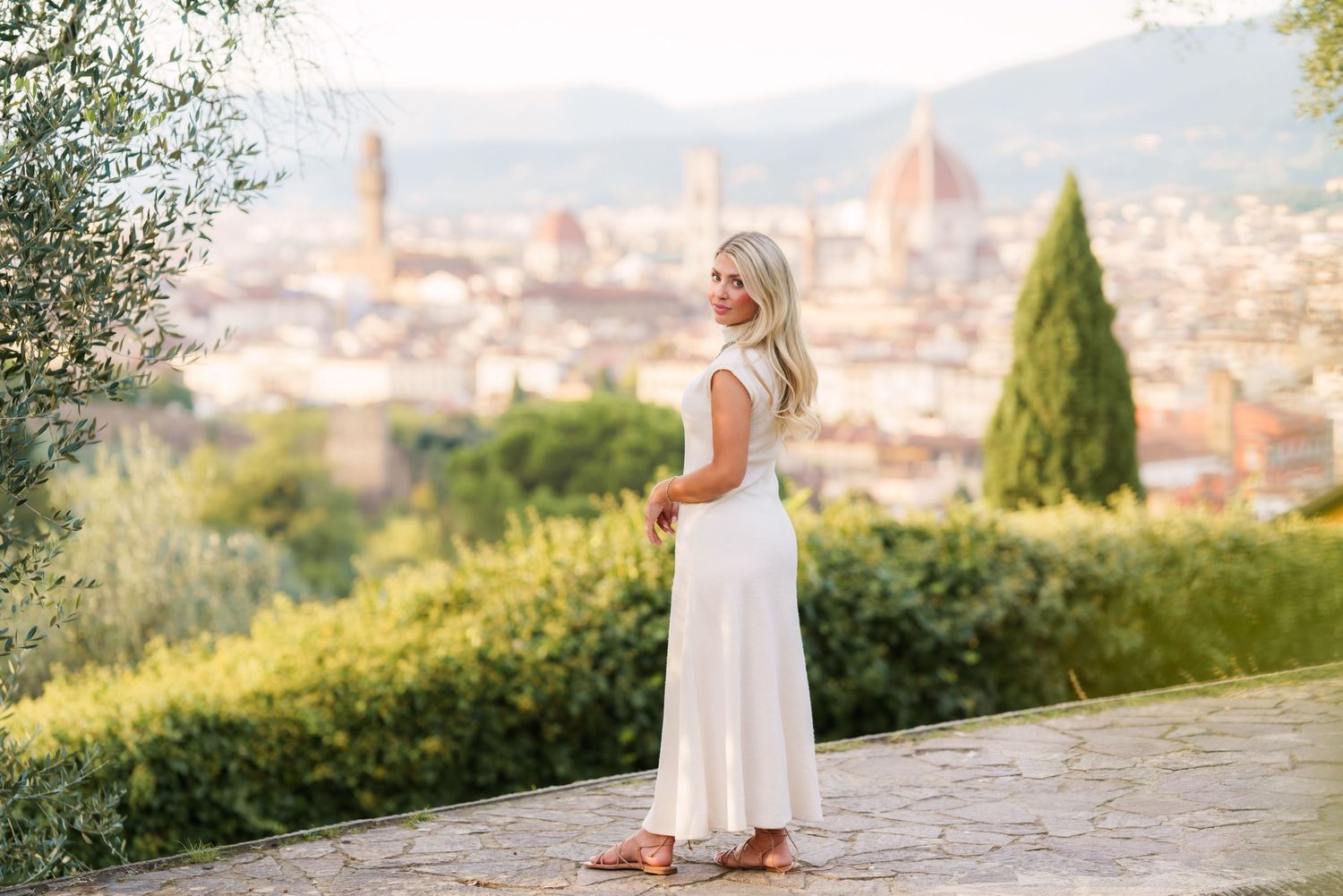 A person in a white dress stands on a scenic overlook with a panoramic view of Florence, Italy in the background.